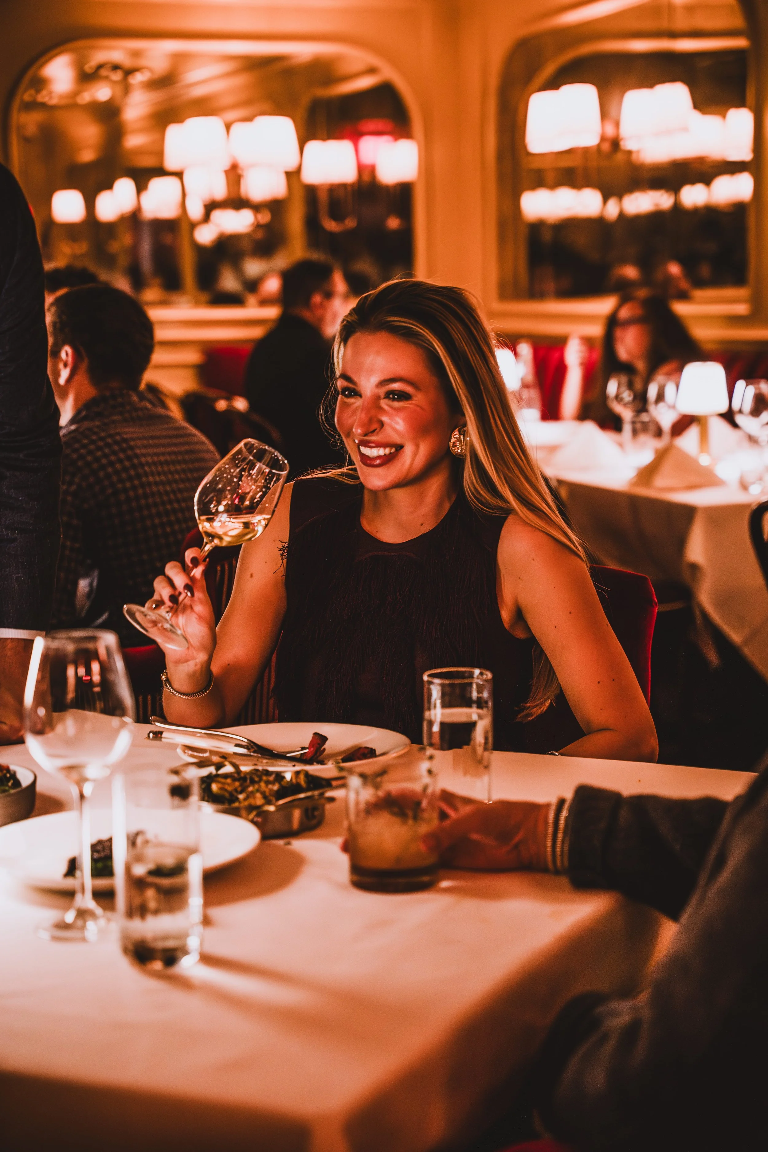 A woman with long blonde hair, smiling and holding a glass of wine, seated at a dinner table in an elegant restaurant with warm lighting and other diners in the background.