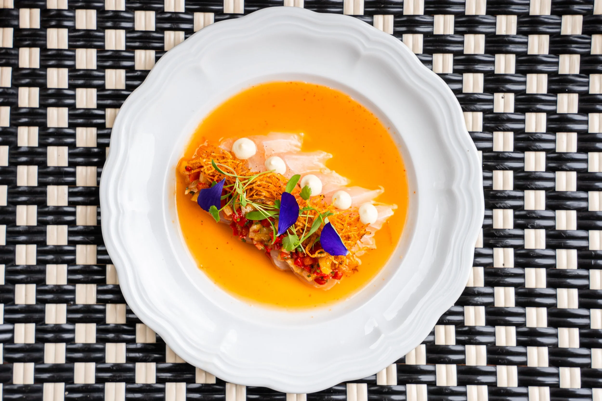 A white decorative plate with a seafood dish featuring fish fillet, orange-colored sauce, garnished with edible flowers, microgreens, and crispy toppings, placed on a black and beige woven placemat.