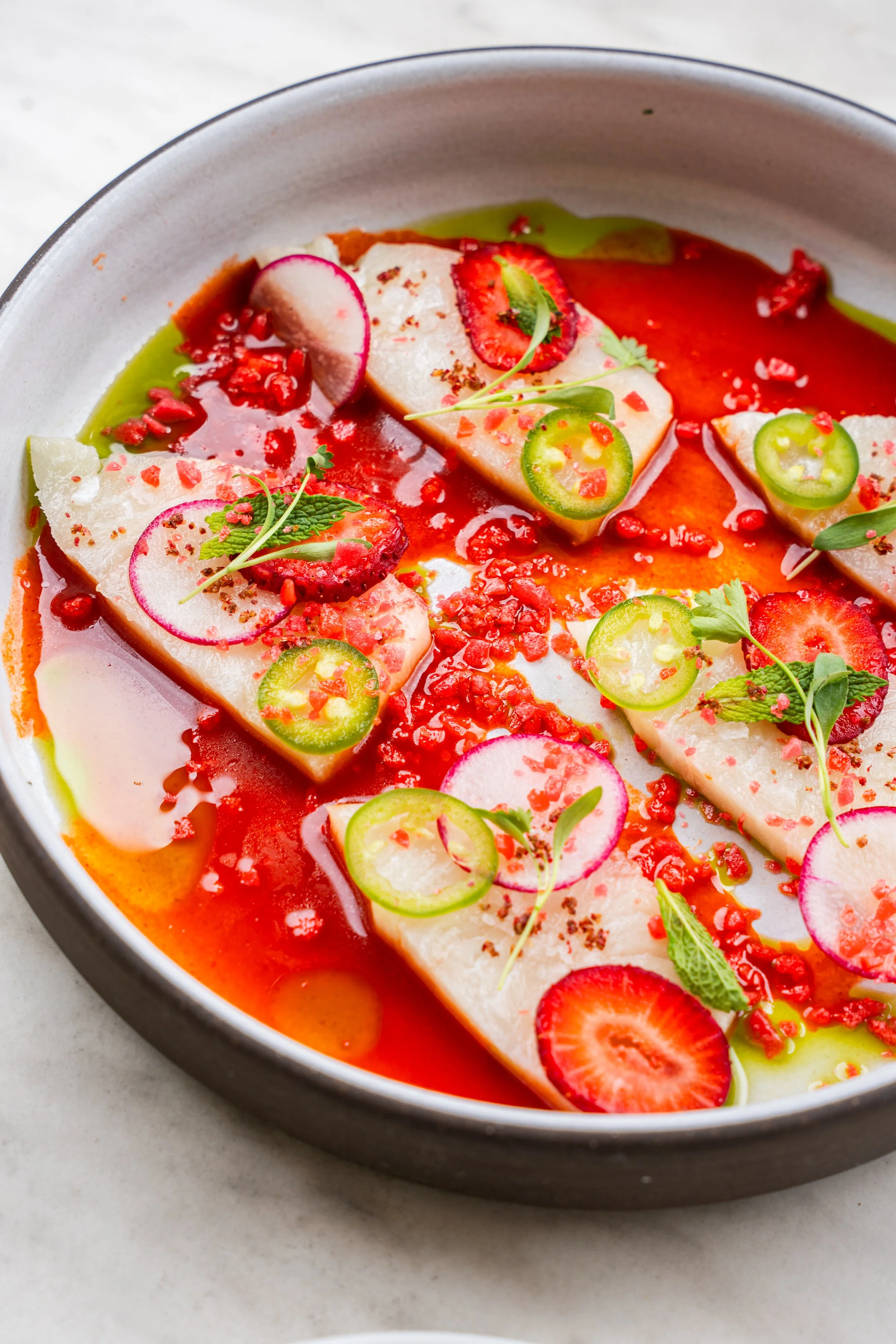 A plate with slices of white fish, topped with sliced strawberries, radishes, green chili peppers, microgreens, and red sauce or seasoning.
