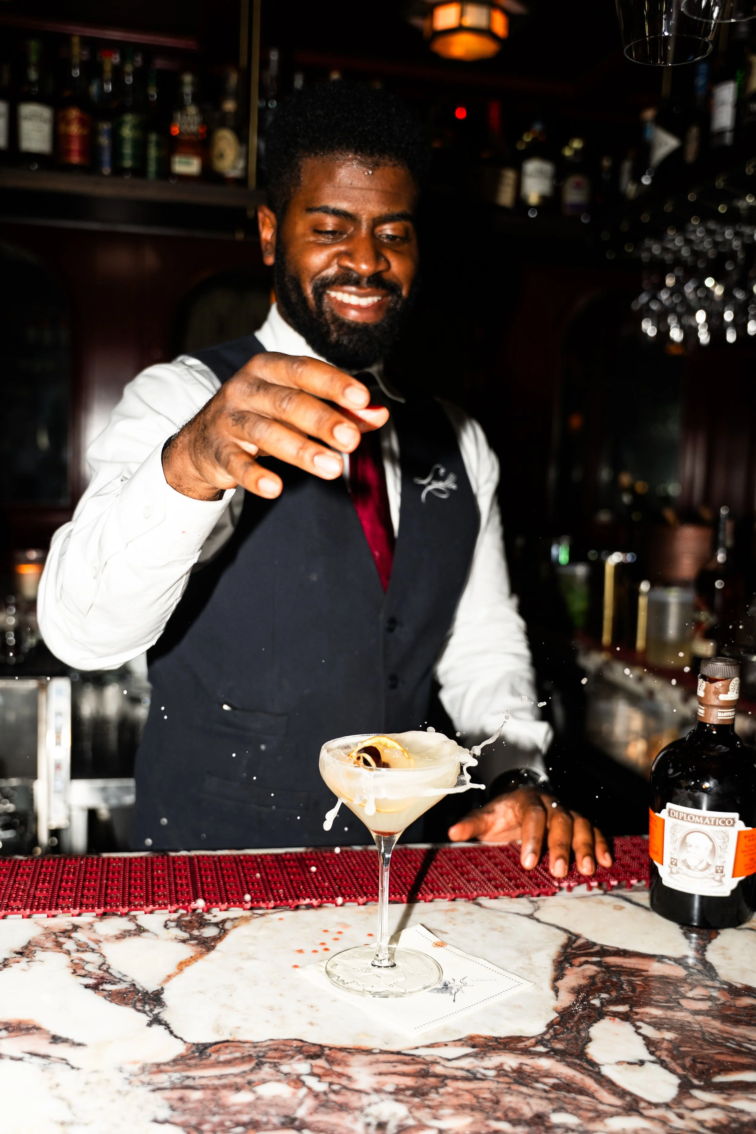 A bartender in a black vest and white shirt is pouring a drink into a cocktail glass, which is splashing foam or cream, in a bar setting with bottles on shelves in the background.
