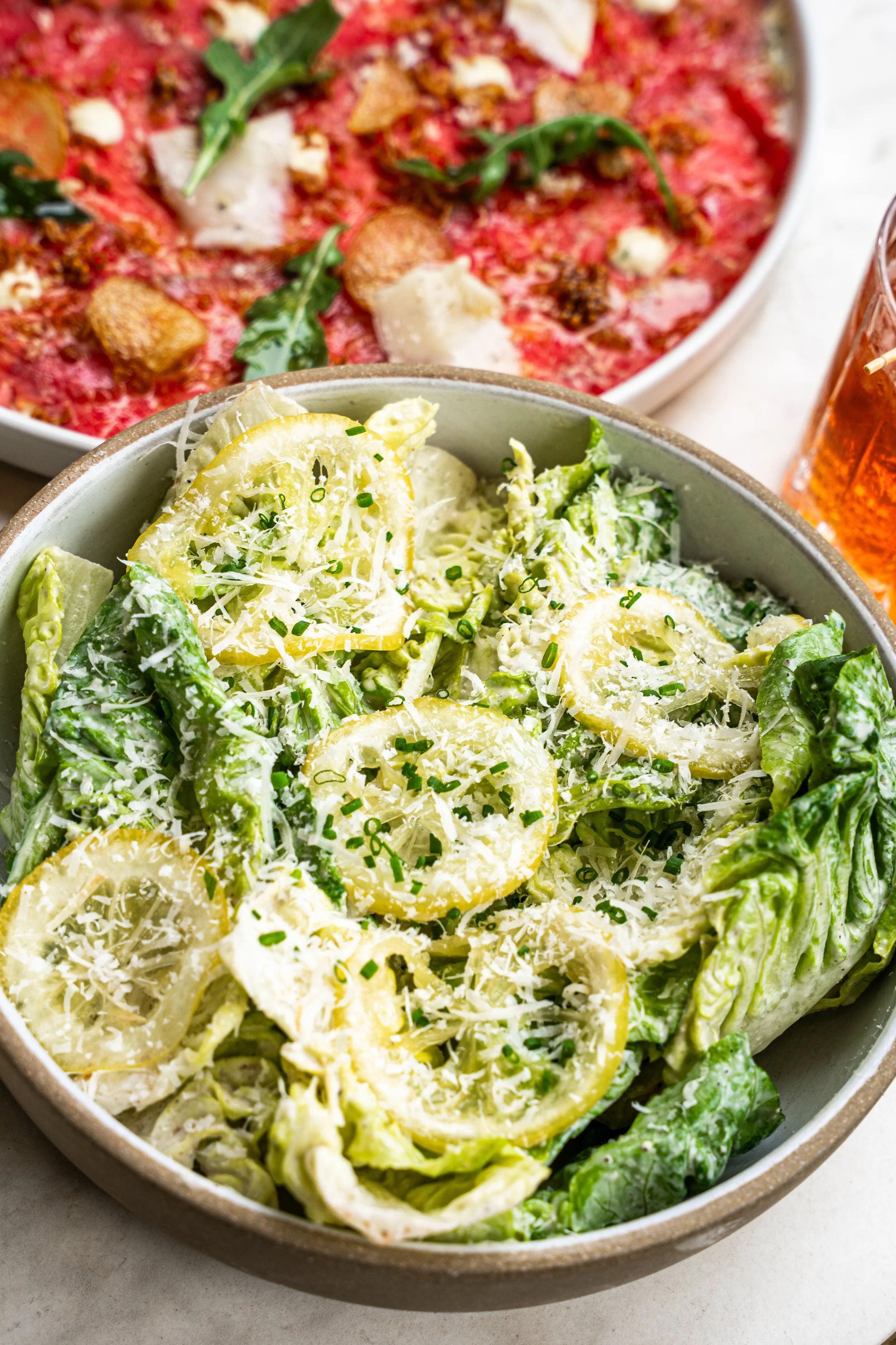 A bowl of Caesar salad with lemon slices, shredded cheese, and chopped green onions.