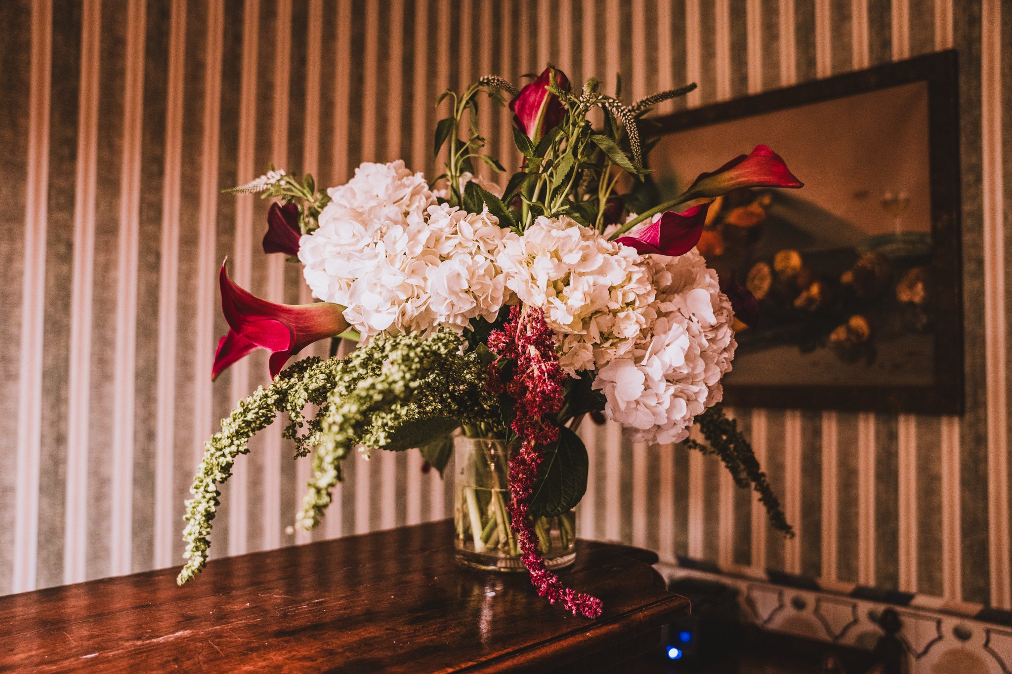 A floral arrangement in a glass vase on a wooden table, featuring white hydrangeas and red calla lilies with greenery, set against a striped wall background.