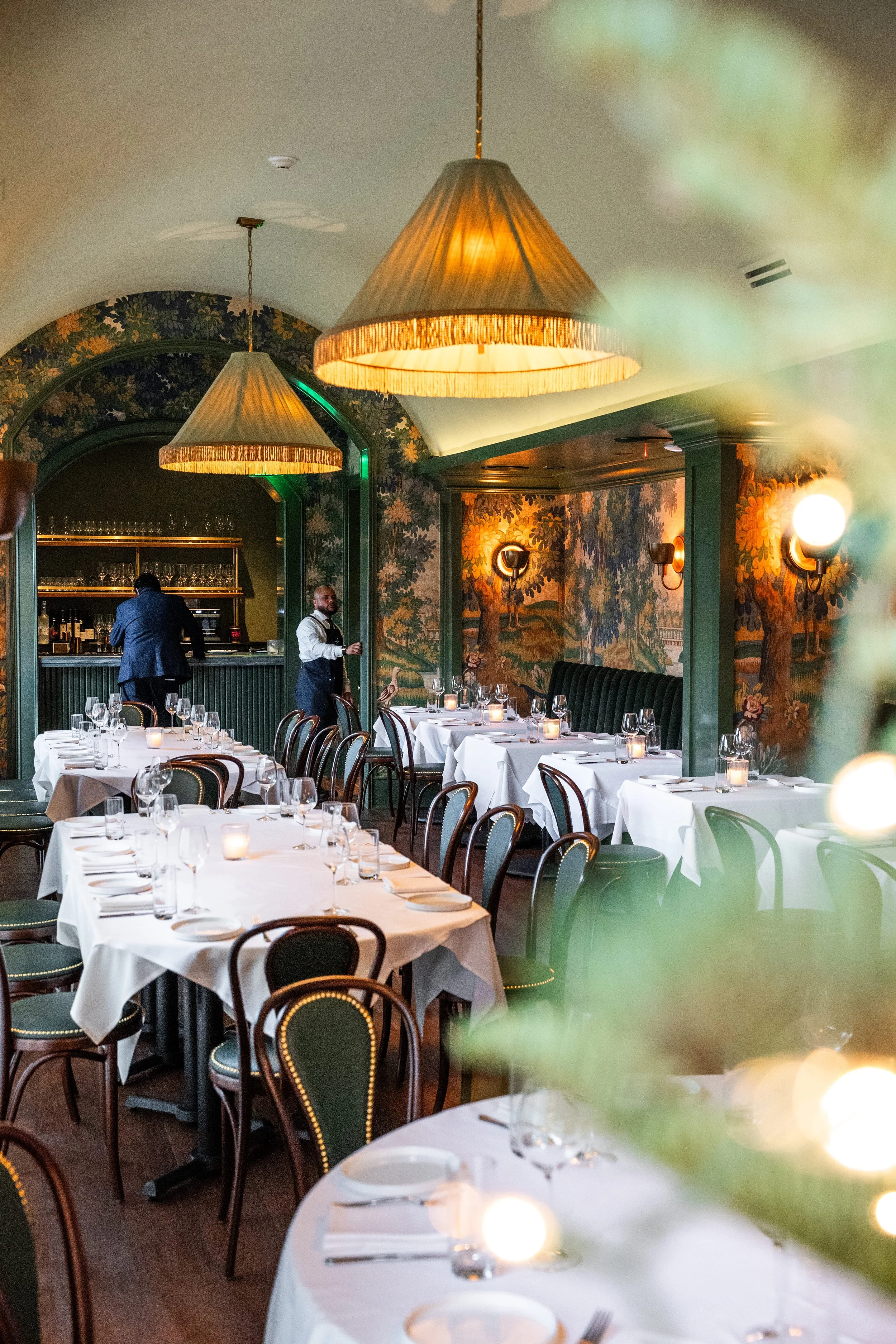 Interior of a restaurant with tables set with white tablecloths, glasses, and candles. Green chairs surround the tables. There are large hanging lamps, artwork of trees and nature scenes on the walls, and two staff members in the background, one working behind the bar and the other attending to the tables.