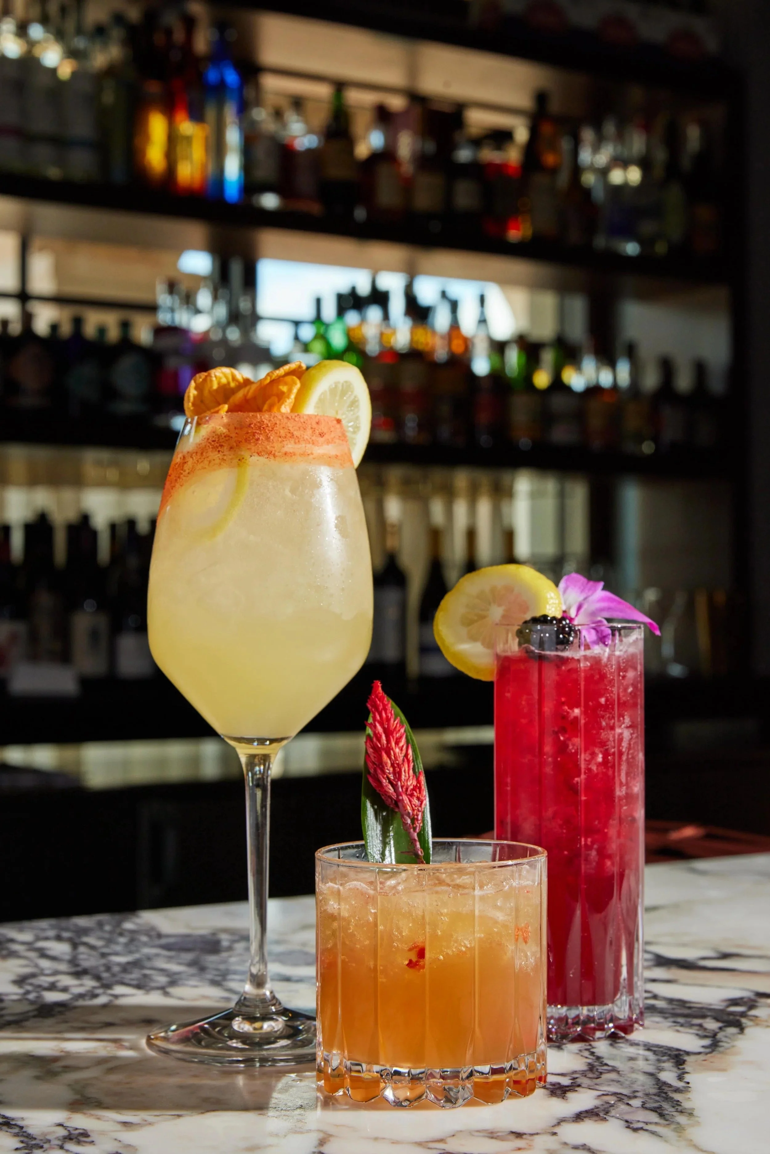 Three colorful cocktails on a marble bar counter with bottles in the background. The first cocktail is a tall, frosty yellow drink garnished with lemon and orange slices. The second is a bright pink beverage decorated with a lemon slice, blueberries,