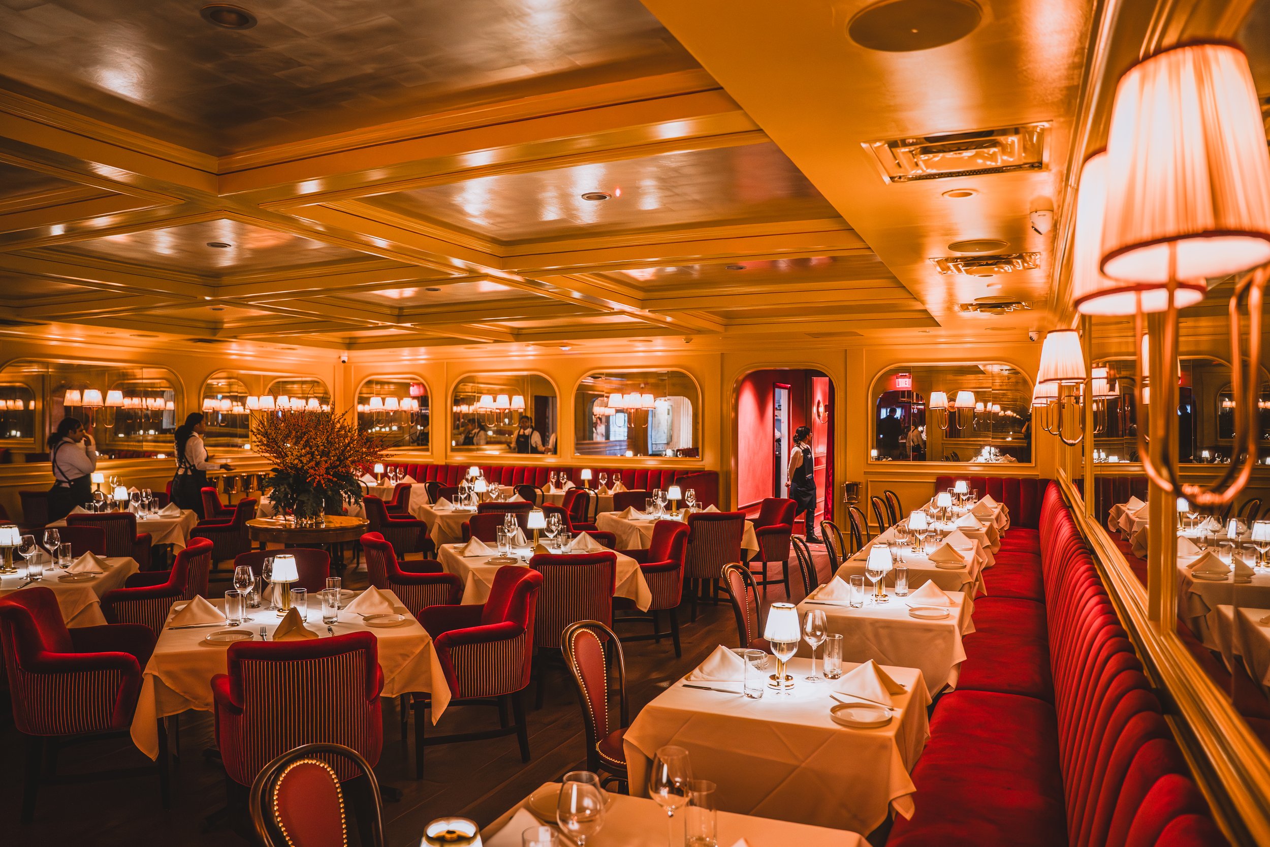 A luxurious restaurant with round tables covered in white tablecloths, elegant red chairs, and soft lighting from lamps. The ceiling has a warm glow, and large mirrors line the walls. Waitstaff are attending to the tables, and floral centerpieces dec