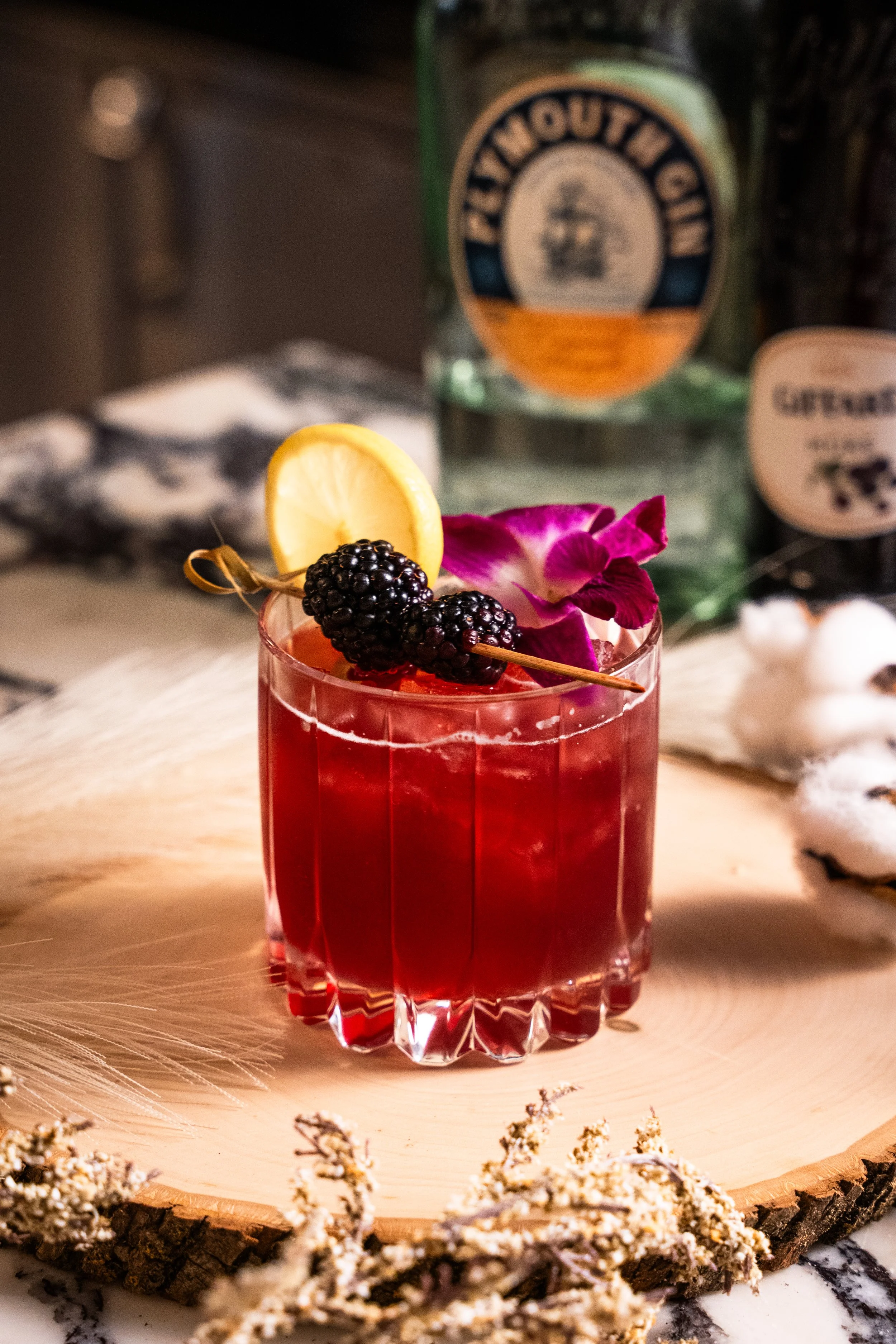 A vibrant red cocktail garnished with blackberries, a lemon wedge, and an orchid flower, placed on a wooden surface with a cut wood slice underneath, with a bottle of Mount Gay Rum and another beverage in the background.