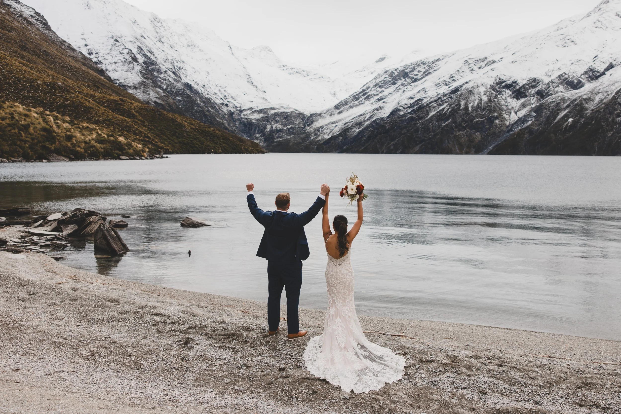 A bride and groom standing at a heli wedding at lachnagar. The groom is dressed in a dark suit, raising his left fist, and the bride in a white wedding dress is holding a wedding bouquet. Elope to Arrowtown in winte, Nicolet Spice wedding celebrant.