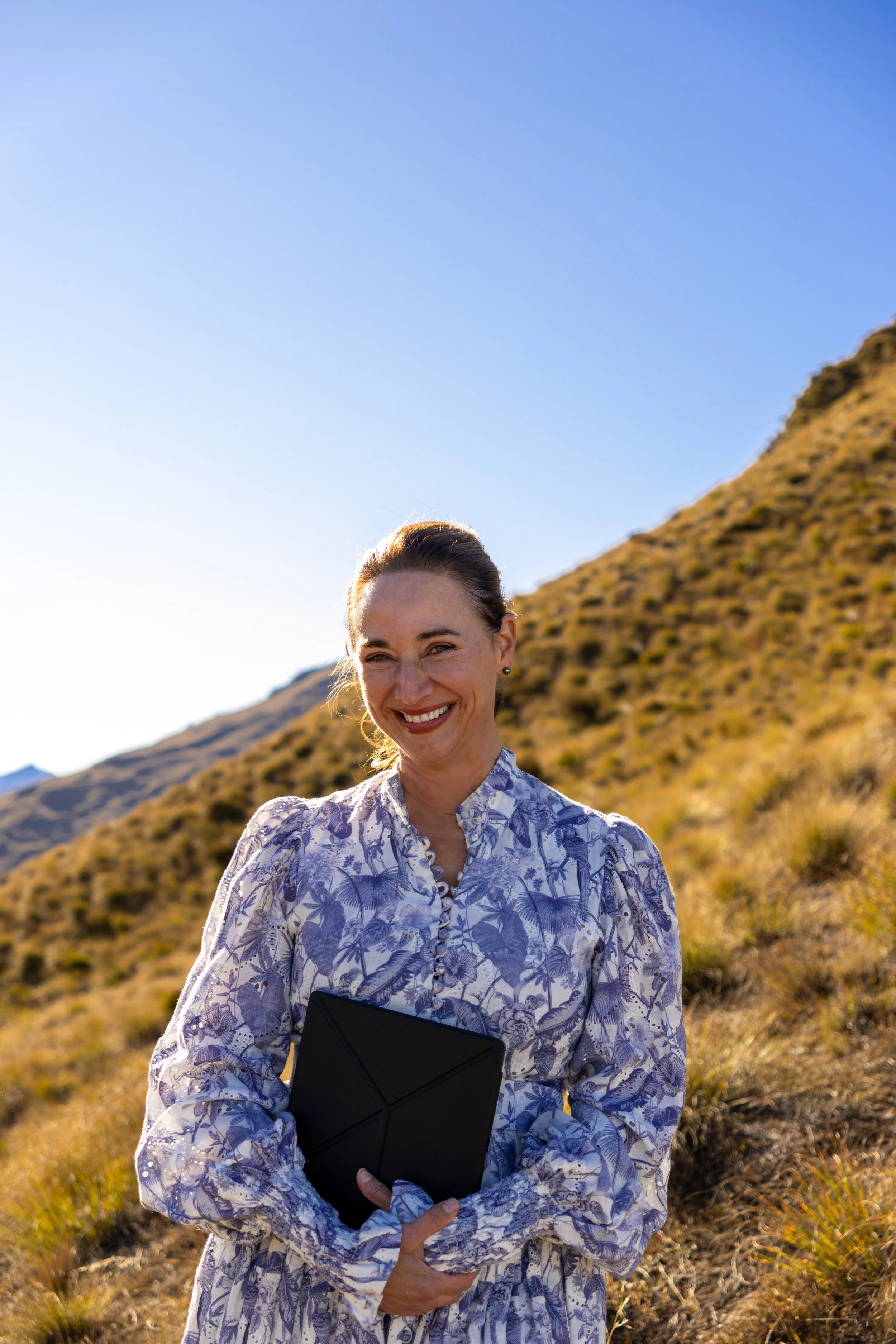 Nicolet Spice smiling and holding a black folder containing wedding vows  stands outdoors on a hillside at a Chinese blessing ceremony with a mountain back ground, under a clear blue sky in beautiful Queenstown.