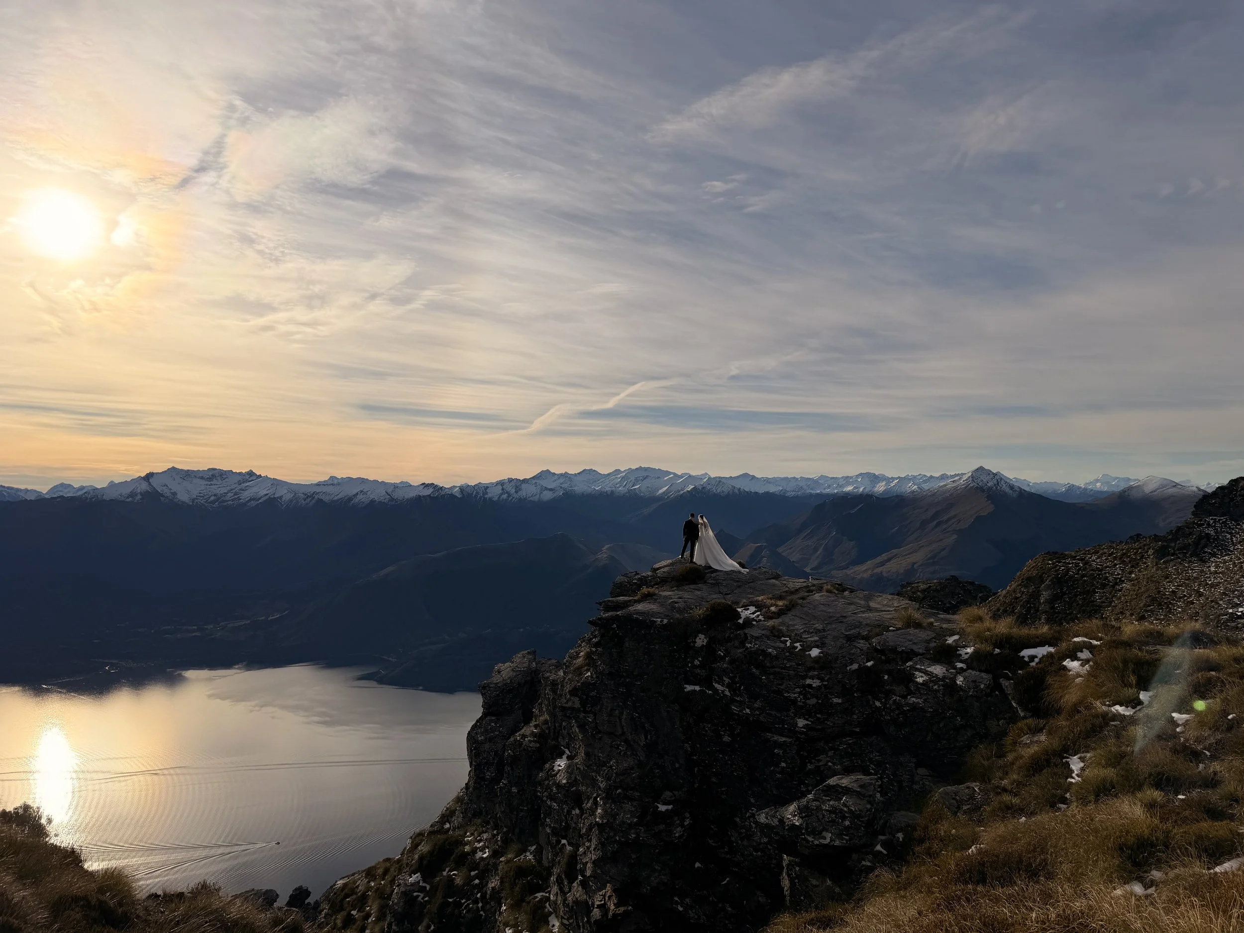 A couple has eloped to Queenstown and held a helicopter wedding standing on a rock ledge overlooking a mountain lake. Sunset wedding , with snow-capped mountains. best location for a heli wedding or elopement
