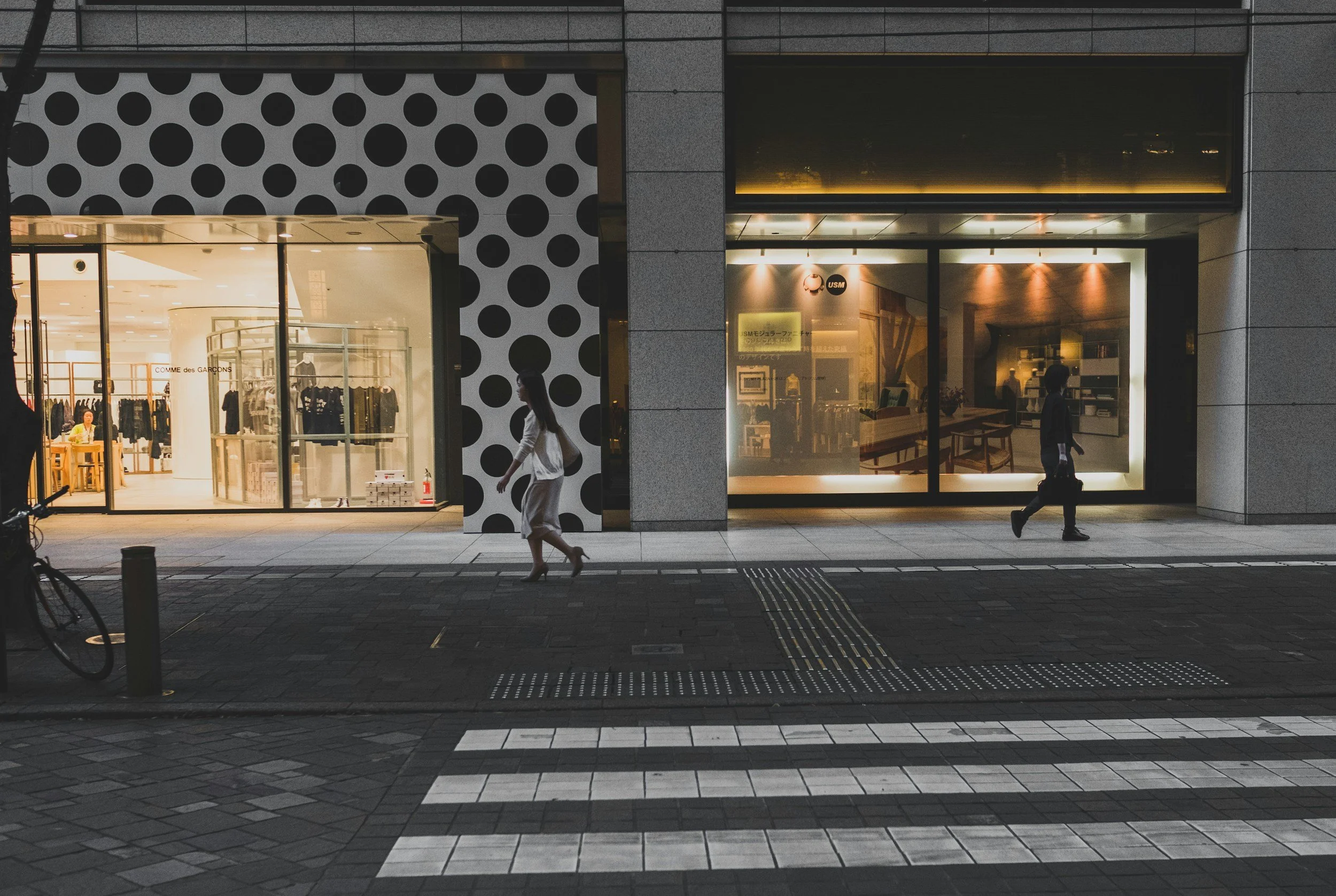 Nighttime street scene showing two pedestrians walking past storefronts with illuminated windows. One woman in a white dress and high heels, and one man in a suit carrying a briefcase. Stores include fashion boutiques, with clothing displays and interior lighting visible.