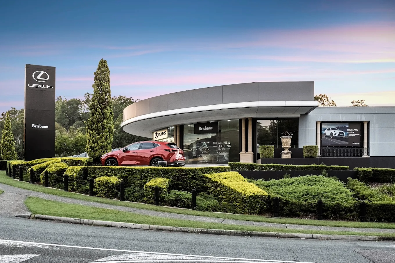 A Lexus car dealership with a red Lexus SUV parked outside, surrounded by manicured greenery, and signage indicating the Brisbane location.
