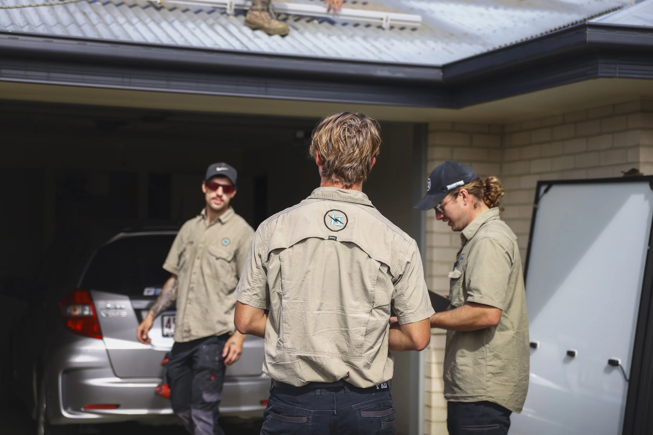 Three men in tan shirts are standing outside a garage, with one woman also in a tan shirt, standing near a silver car. The woman is looking at her phone, and the man in the middle has his back to the camera. The garage has a brick wall and a white door.