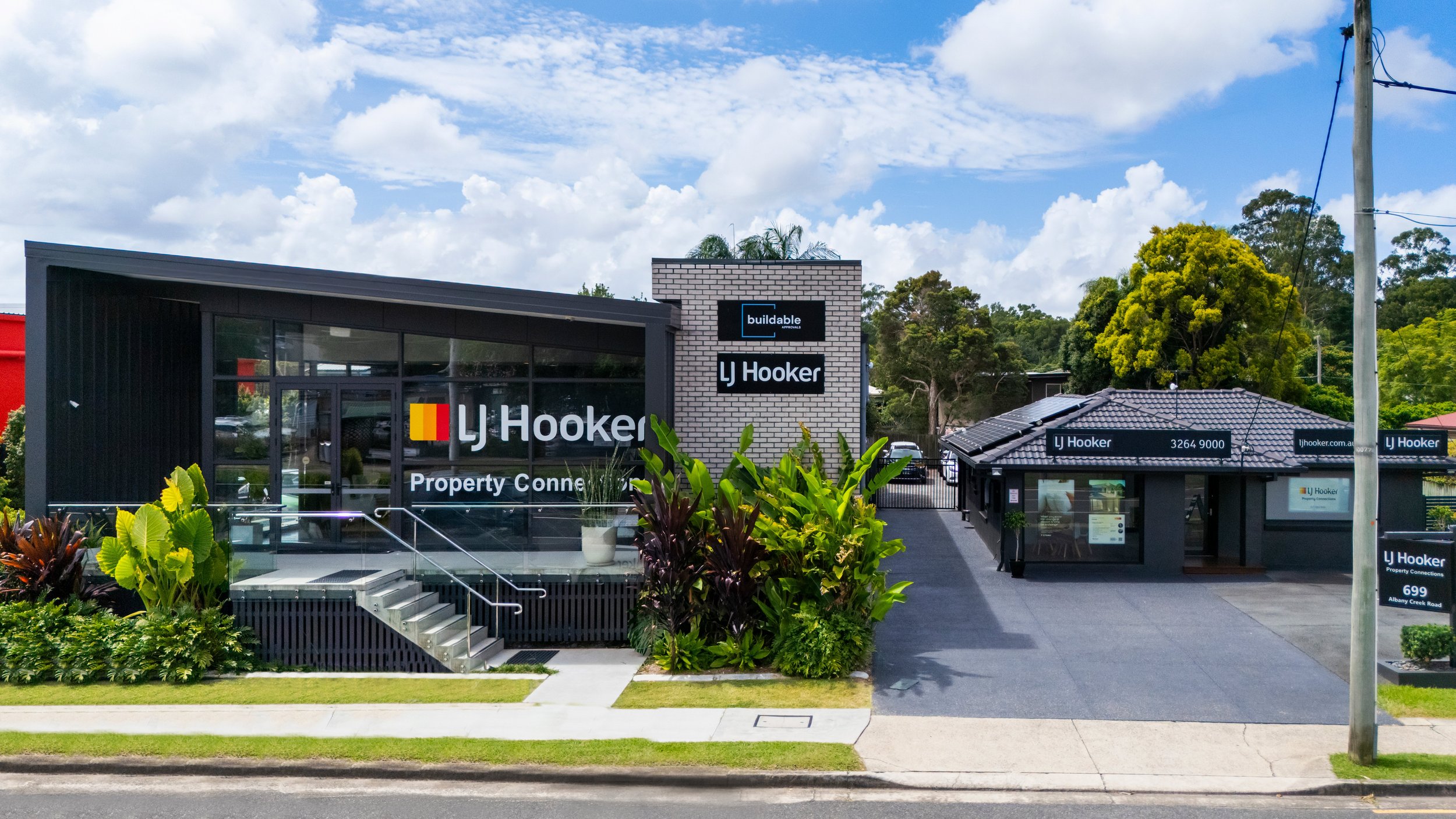 LJ Hooker real estate office with a black modern building on the left and a small office on the right, surrounded by greenery, under a blue sky with clouds.