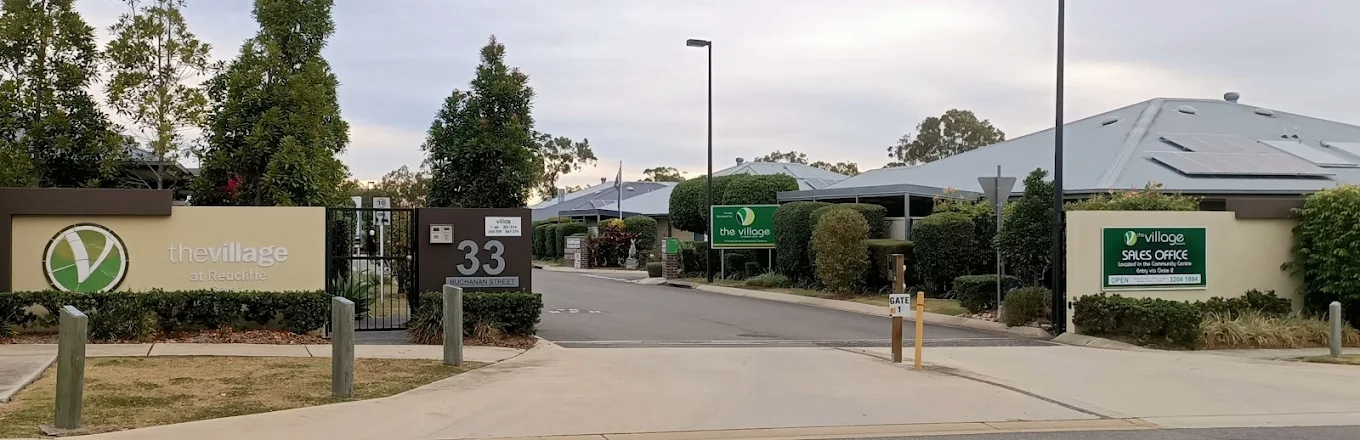 Entrance to The Village at Redcliffe, showing signs for the village and sales office, along with a gated entry, trees, and a building with a metal roof in the background.