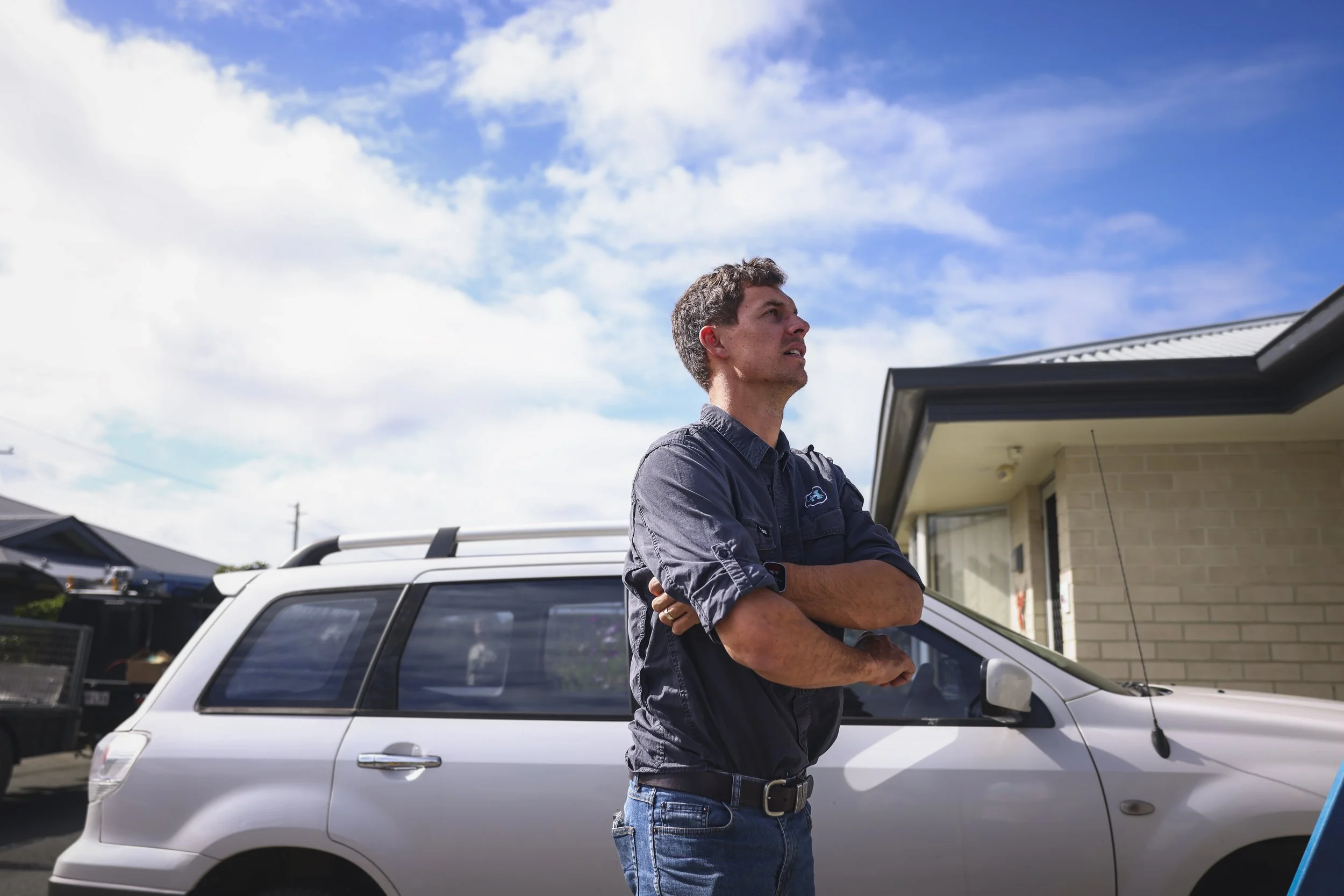 A young man stands outdoors with arms crossed, looking up. He is near a silver car and a house, under a partly cloudy sky.