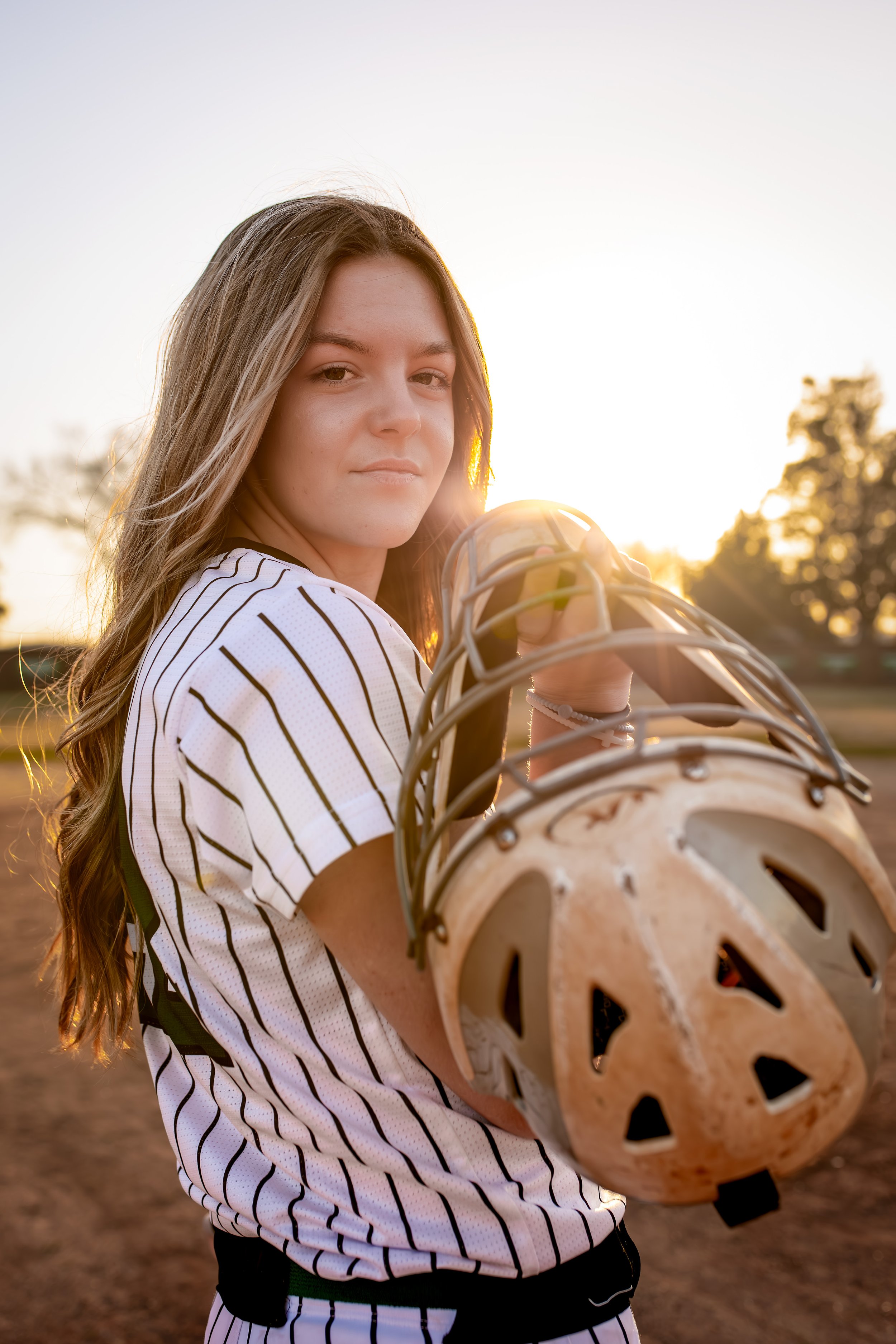 A young woman in a baseball jersey holding a helmet on a baseball field during sunset.