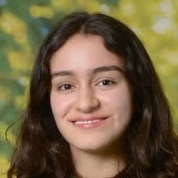 A young woman with long, wavy brown hair smiling at the camera, standing outdoors with a blurry background of yellow and green foliage.