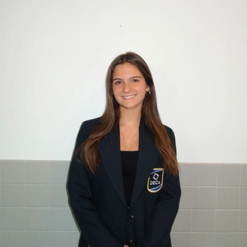 A young woman with long brown hair, smiling, wearing a navy blazer with a 'DECA' badge, standing against a plain white and tiled wall.