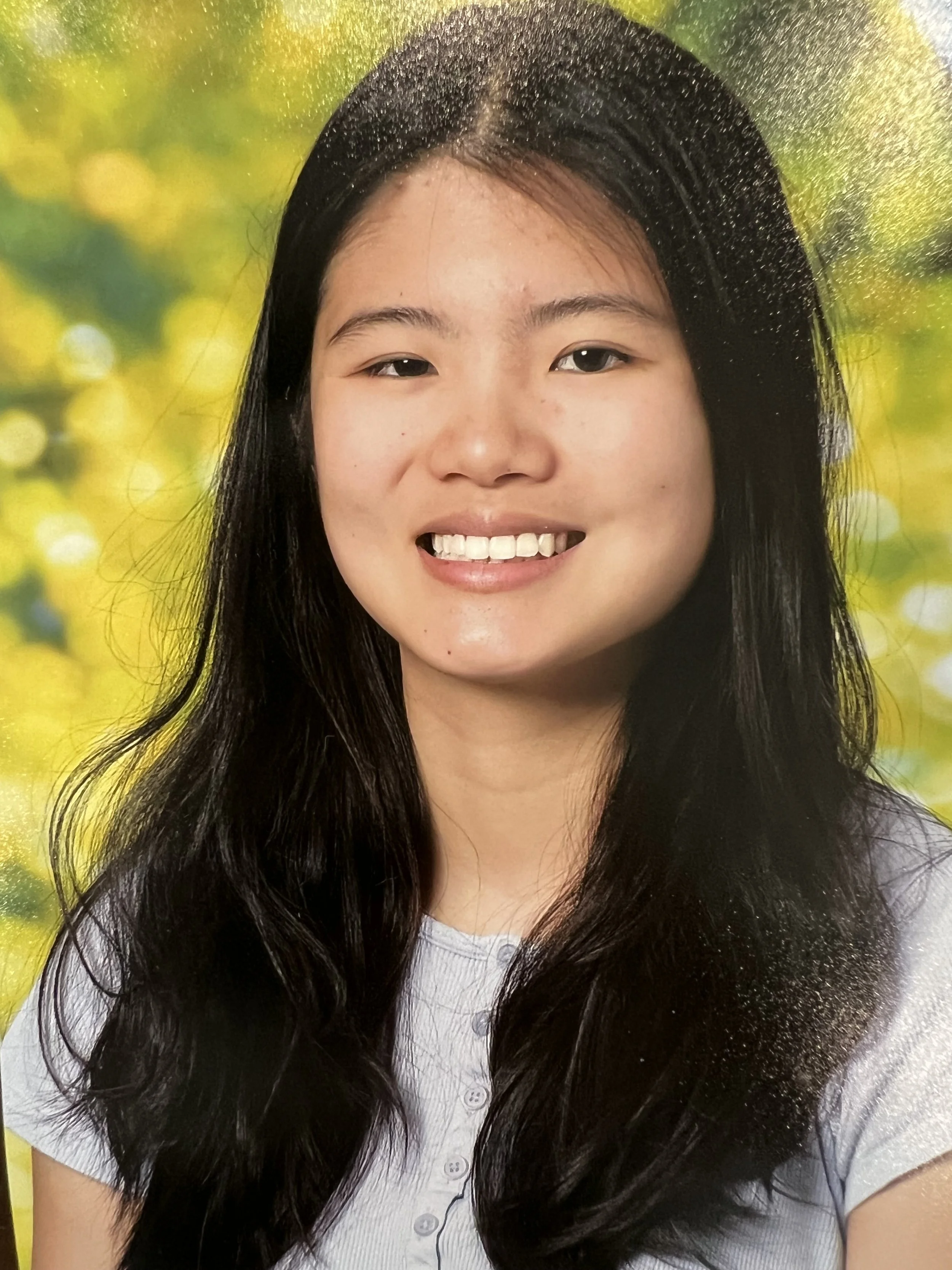 A young woman with long black hair smiling in front of a blurred yellow and green background.