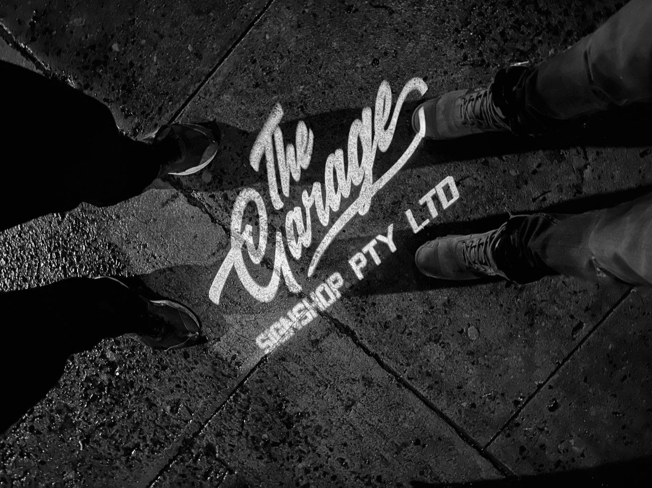 Black and white photo of two people's legs and feet standing on a wet sidewalk with a shadow cast.