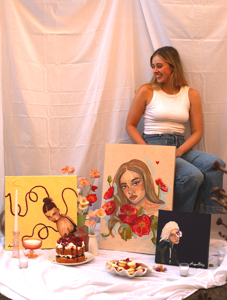 A woman sitting next to a display of colorful paintings, flowers, and desserts.