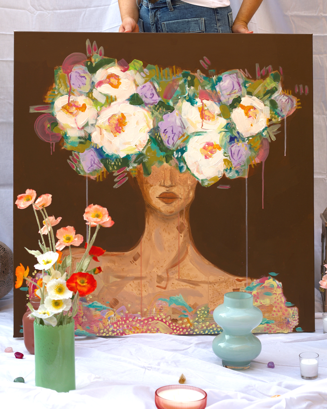An art display featuring a painting of a woman with flowers as hair, surrounded by flower arrangements and decorative vases on a white table.