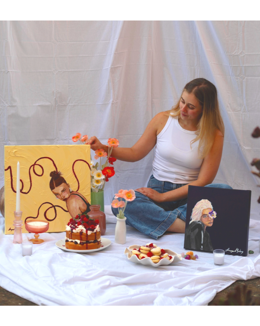 A woman sitting on a white cloth surrounded by artwork, candles, a cake with strawberries, and flowers, arranging flowers in a vase. Two paintings are visible, one of a woman with her hair in a bun and tentacles, and another of a woman with white hai