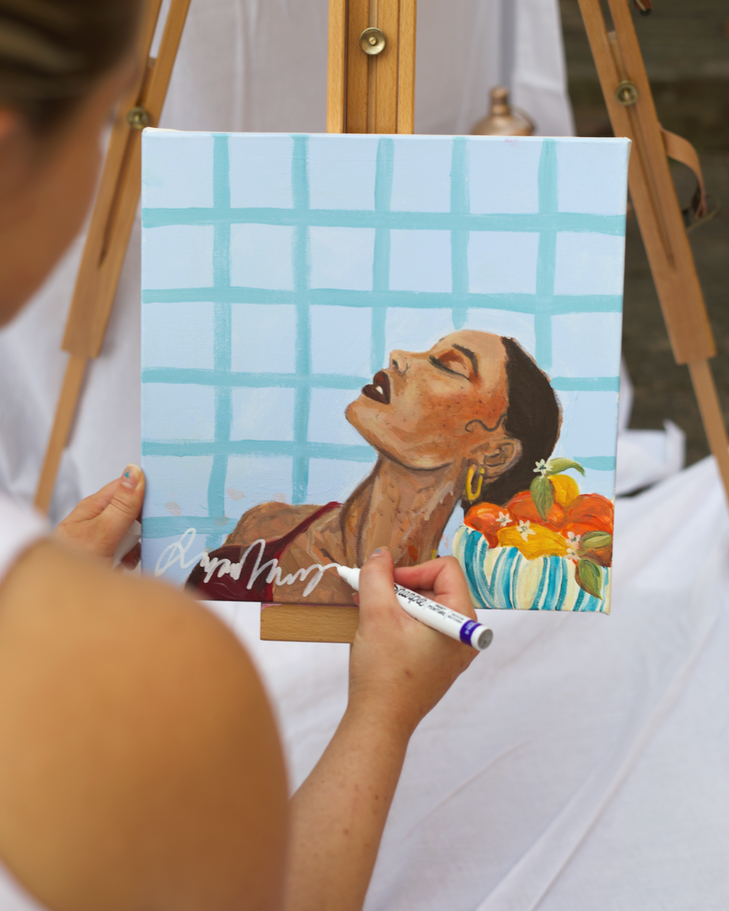 Person painting a portrait of a woman with dark hair and earrings, with a patterned background and a bowl of oranges nearby.