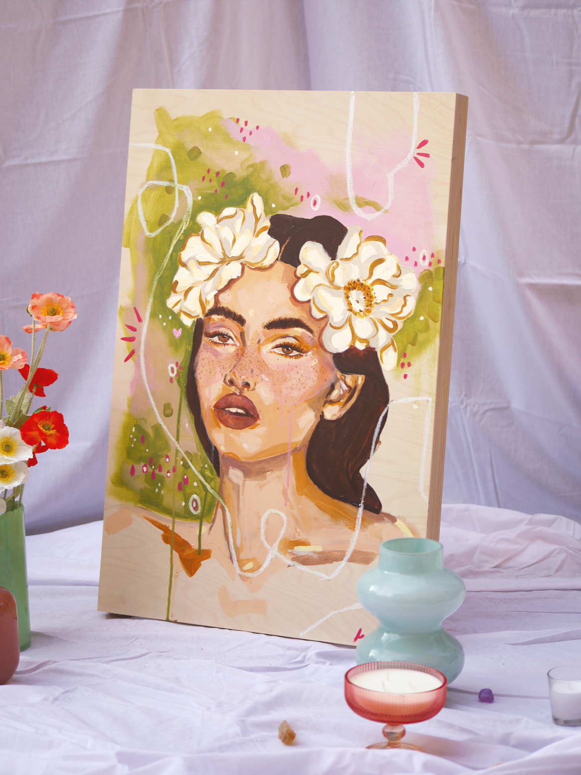 Colorful painted portrait of a woman with freckles, dark hair, and a flower crown on a wooden canvas. The artwork is surrounded by various decorative vases, flowers, and candles on a white surface.