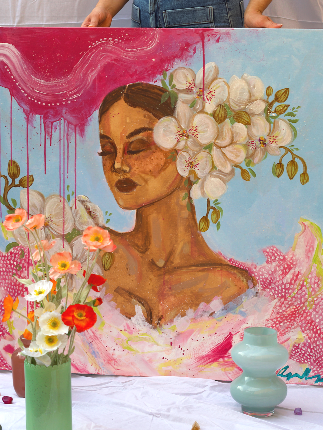 A colorful painting of a woman with closed eyes, dark hair, and freckles, surrounded by white and pink flowers, with a pink and blue background. In front of the painting, there is a vase with orange, white, and pink flowers and a light green ceramic 