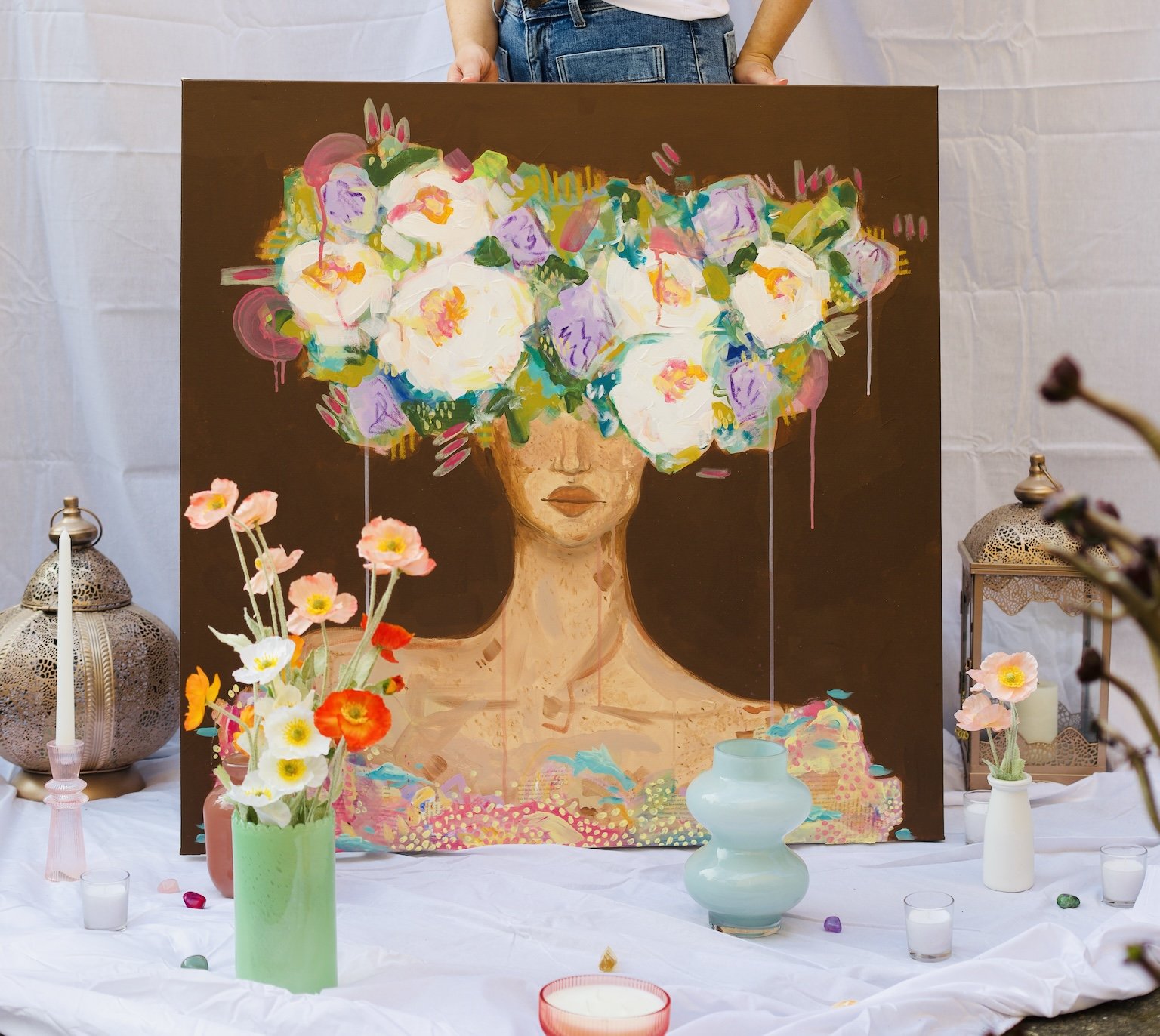 A colorful painting of a woman's face with her hair made of large, vibrant flowers. The painting is displayed on a table with various flower vases, candles, and decorative lanterns around it.