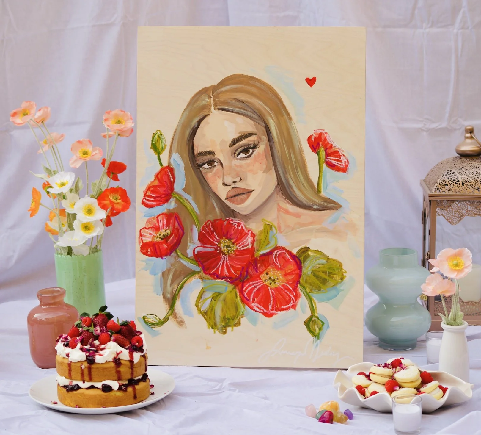 A colorful painting of a woman's face surrounded by red poppies, displayed on an easel. The setup includes flowers in a green vase, pink and peach vases, a layered berry and cream cake, a plate of heart-shaped cookies with berries, and decorative can