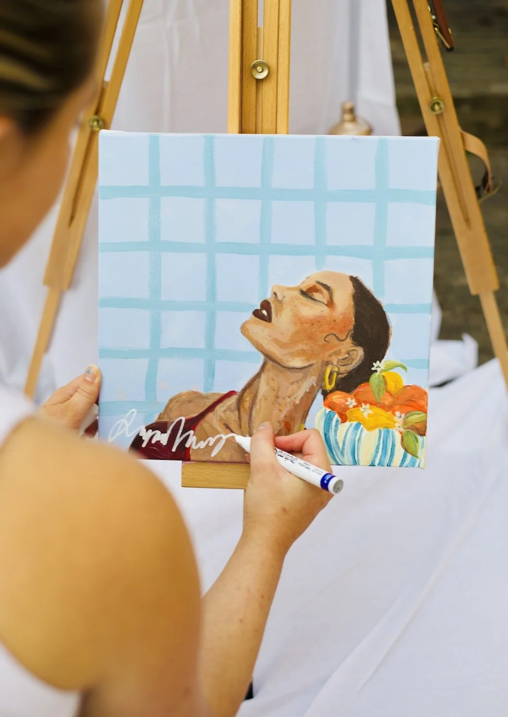 Person creating a portrait painting of a woman with closed eyes, wearing a red top and gold earrings, with a basket of flowers nearby, in front of an easel outdoors.