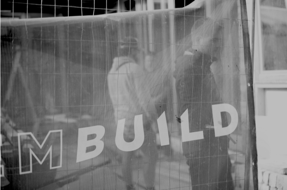 Reflection of two people working on building a project behind a transparent plastic sheet with the word 'BUILD' on it.