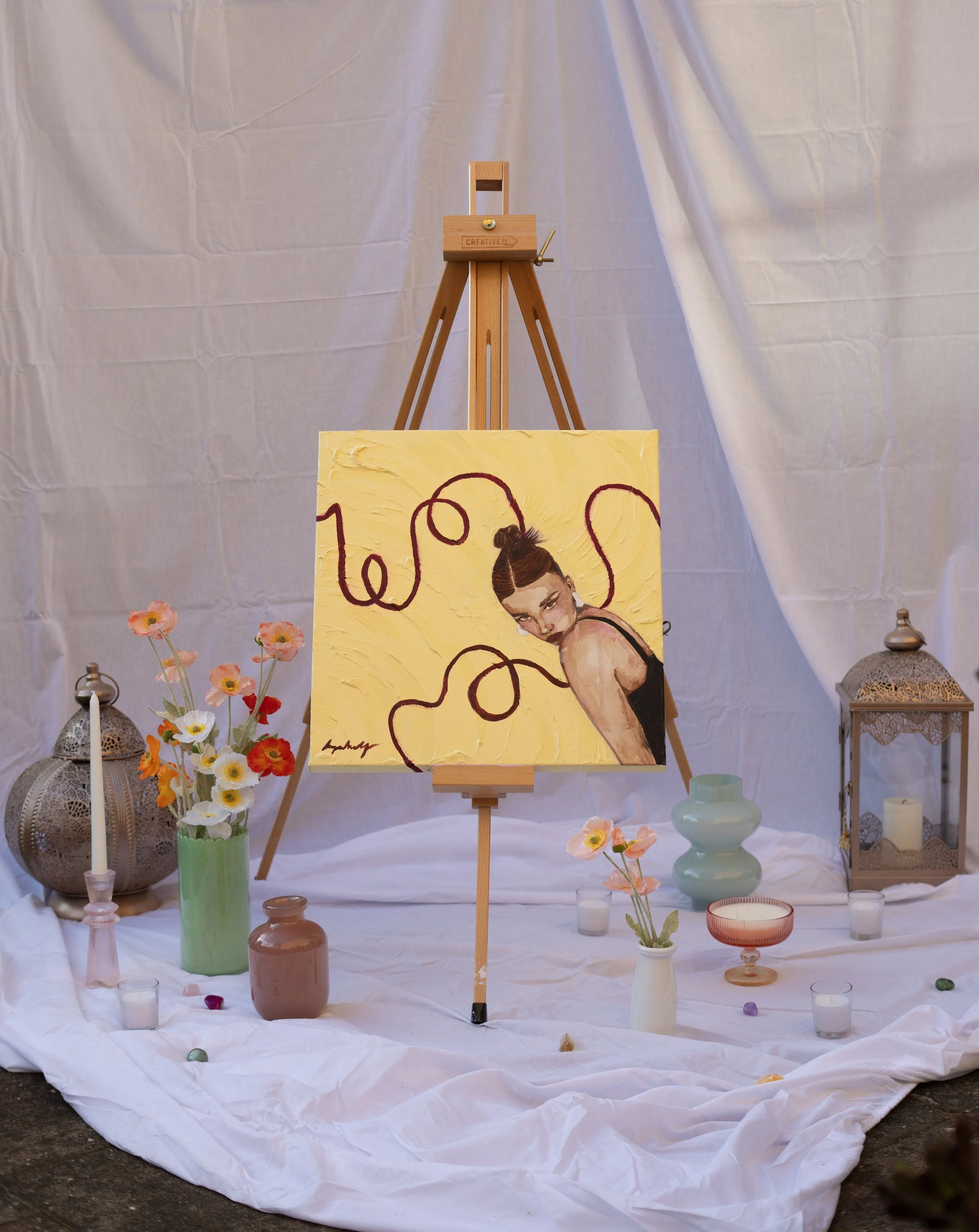 A painting of a woman with a bun hairstyle on a yellow background with abstract lines, displayed on an easel, surrounded by vases with flowers, candles, and decorative lanterns on white fabric.
