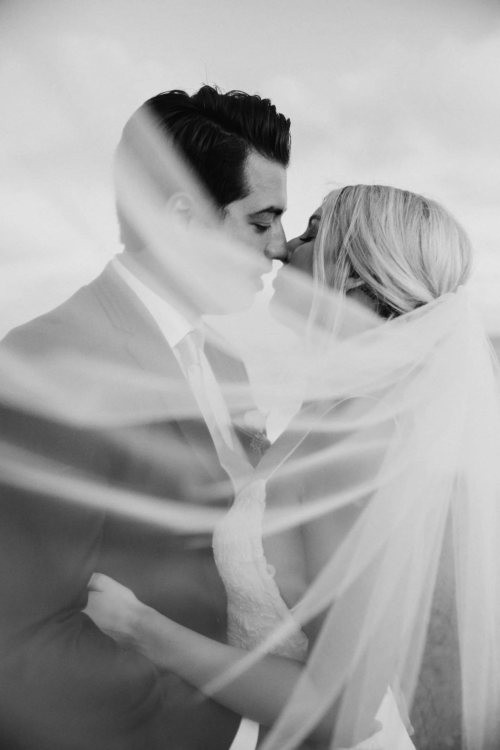 Black and white photo of a bride and groom kissing, with a sheer veil partially covering them.
