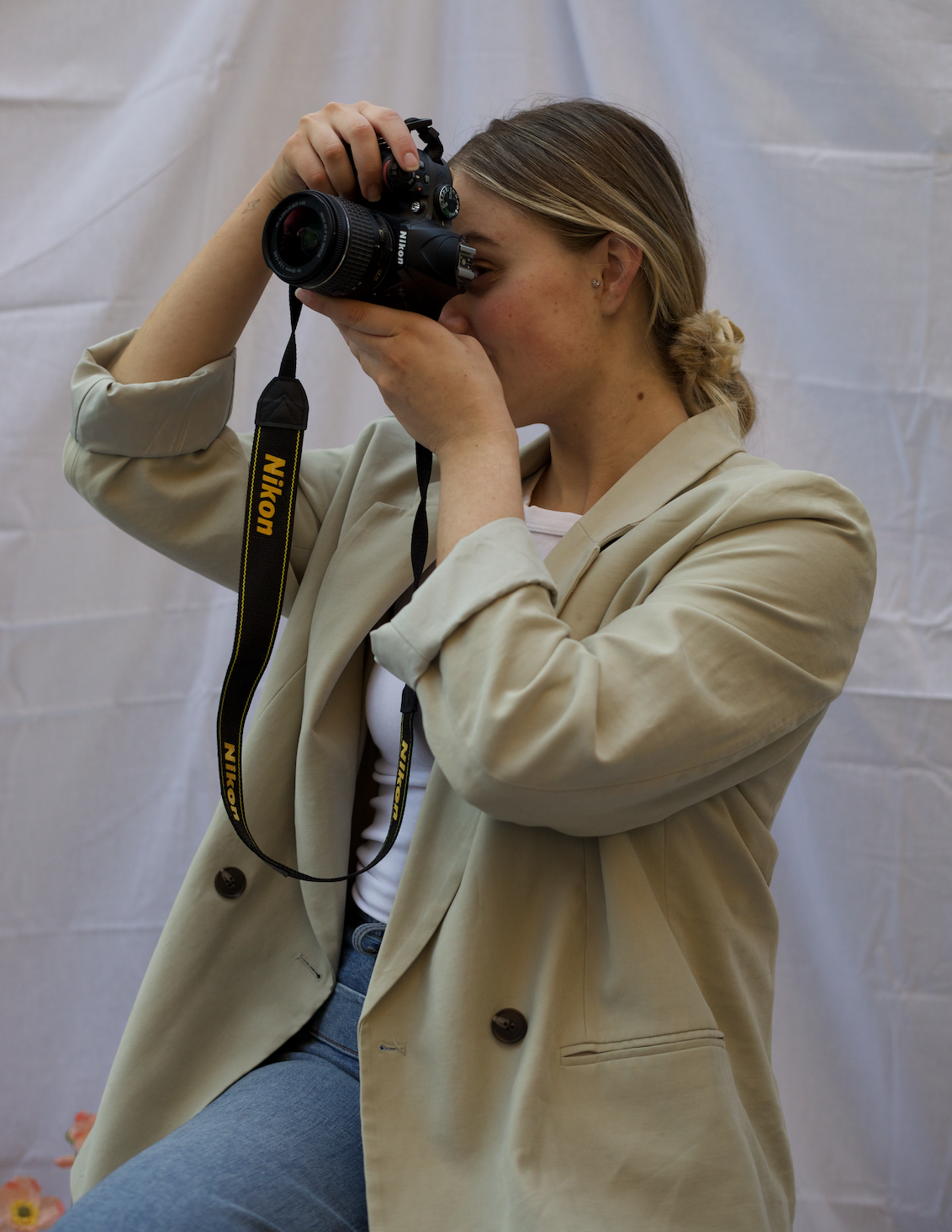 A woman with light brown hair tied in a bun, wearing a beige blazer and white shirt, is taking a photo with a Nikon camera.