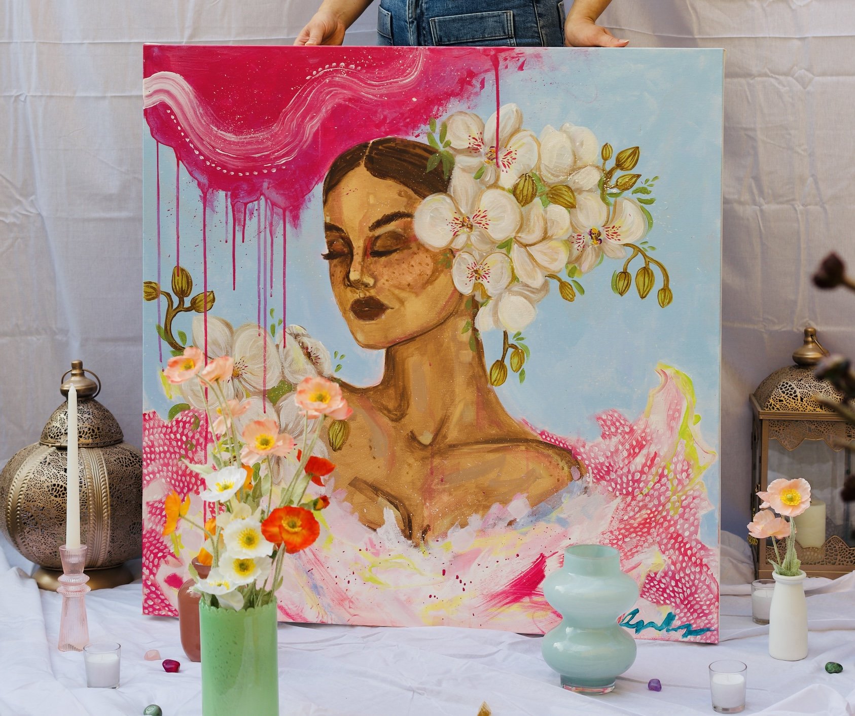 A colorful painting of a woman with closed eyes and dark lipstick, decorated with white flowers and surrounded by pink, blue, and abstract elements, displayed on an easel with floral arrangements and decorative lanterns around.