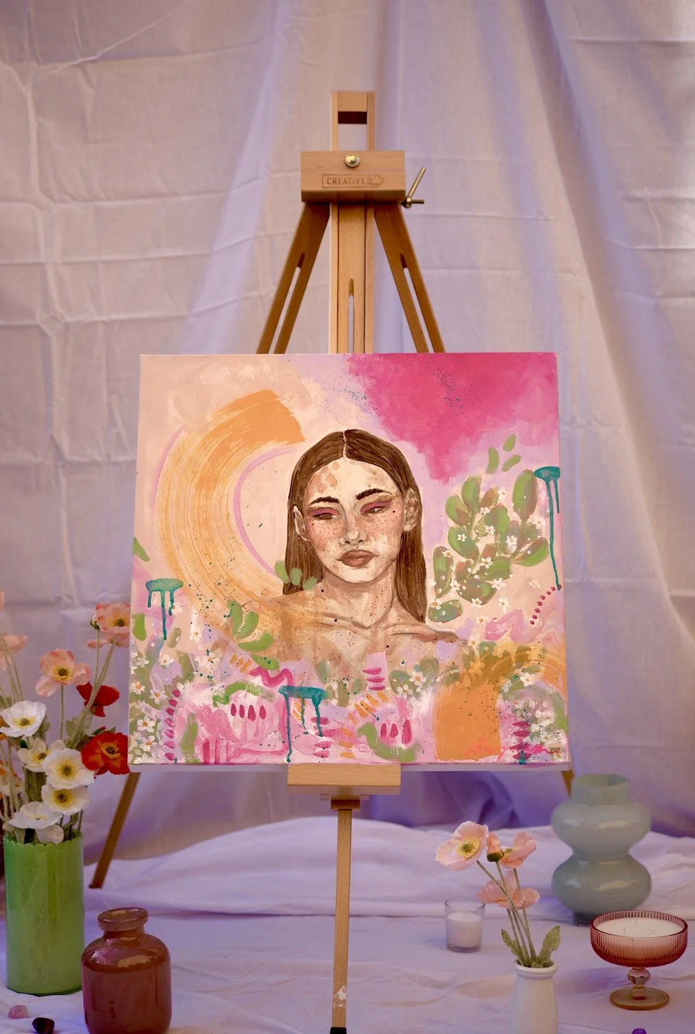 A colorful painting of a woman with long brown hair, surrounded by abstract pink, orange, and green shapes, on a wooden easel with flowers and vases in the background.