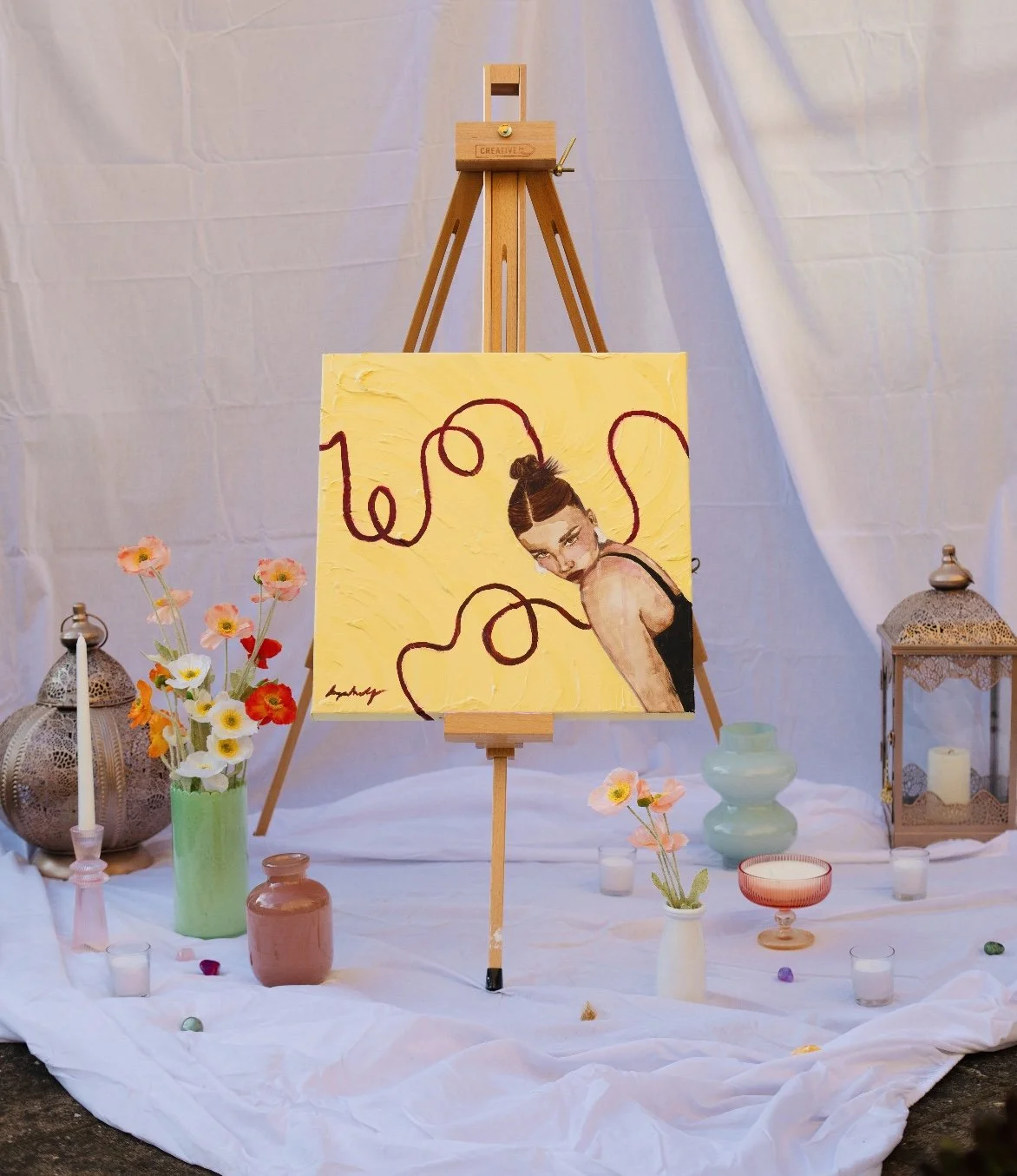 An art display featuring a painting of a woman. The painting background is yellow with red swirling lines, and the woman is looking over her shoulder. There are vases with flowers, candles, and decorative lanterns arranged on the white fabric beneath