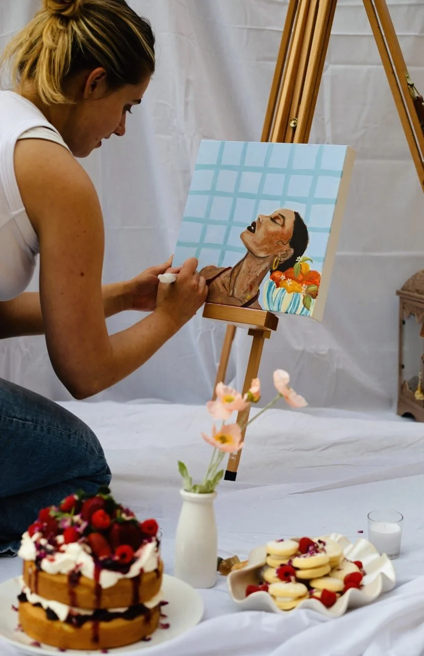 A woman painting a portrait of a woman with dark hair and earrings on a canvas on an easel, with floral decorations and snacks on a white surface in front.