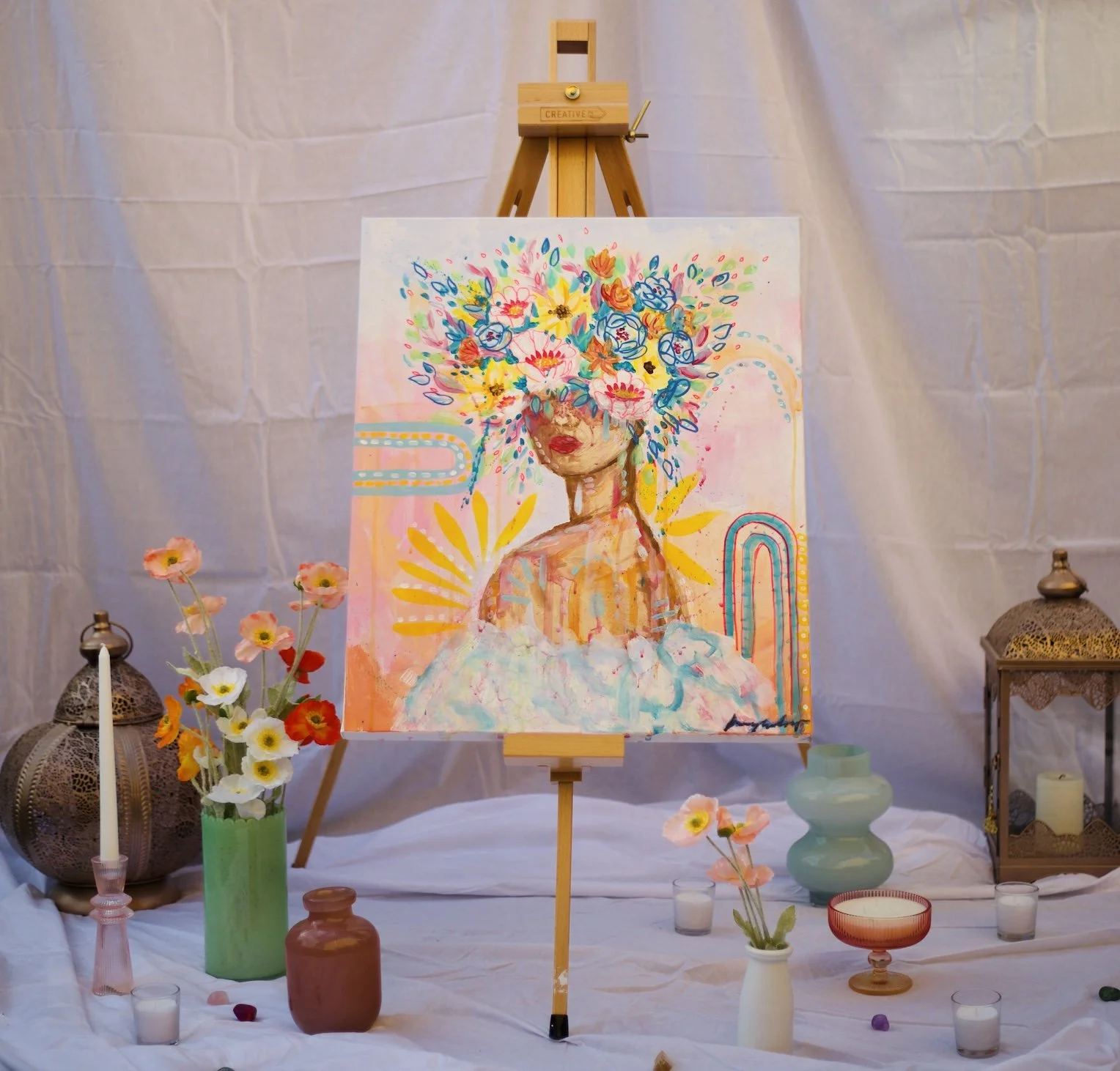 A colorful abstract acrylic painting of a woman with flowers covering her face on an easel, surrounded by candles, vases with flowers, and decorative lanterns on a white cloth background.