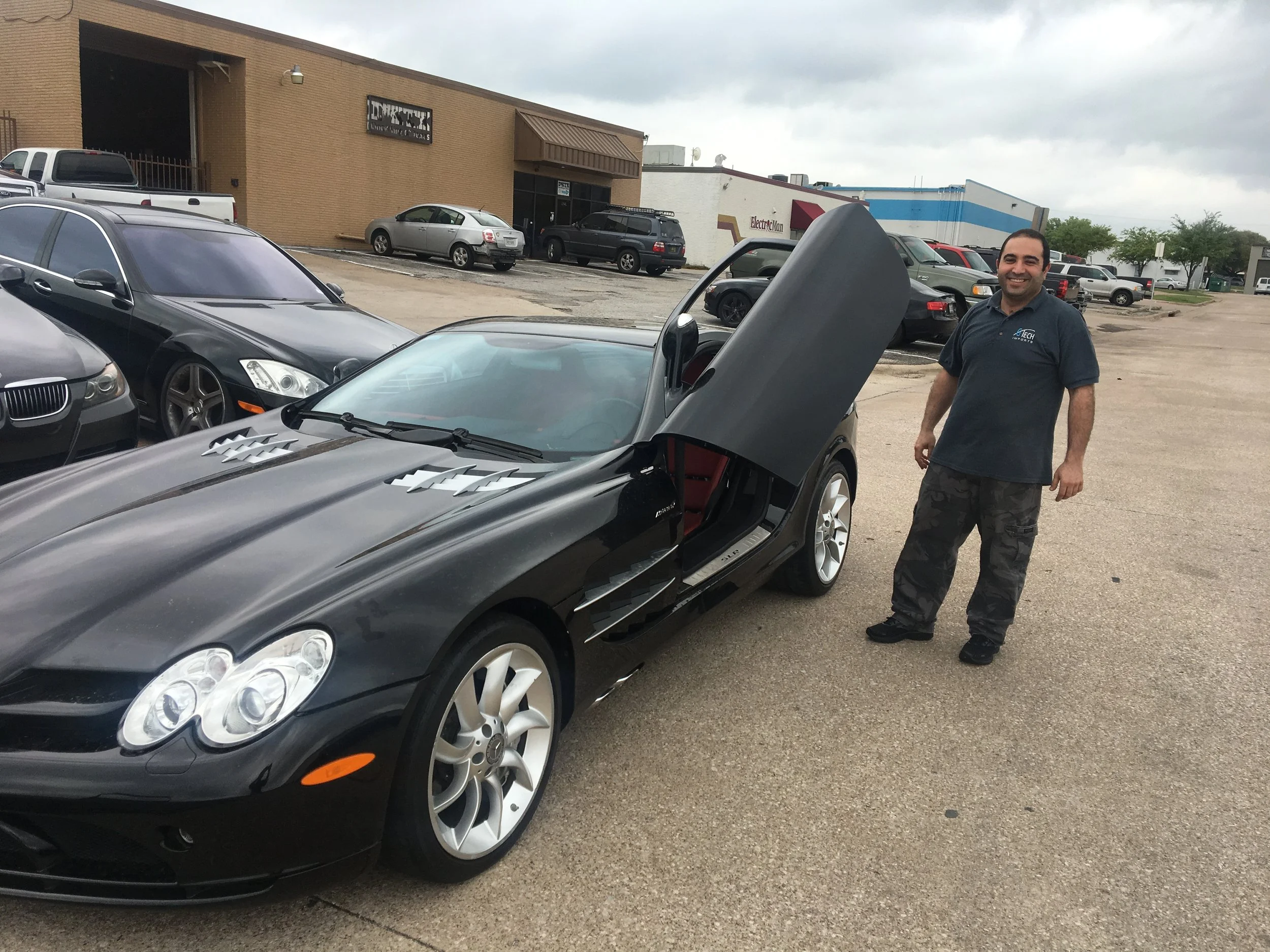 Man standing next to a black sports car with gullwing door open in a parking lot with other cars and a building in the background.