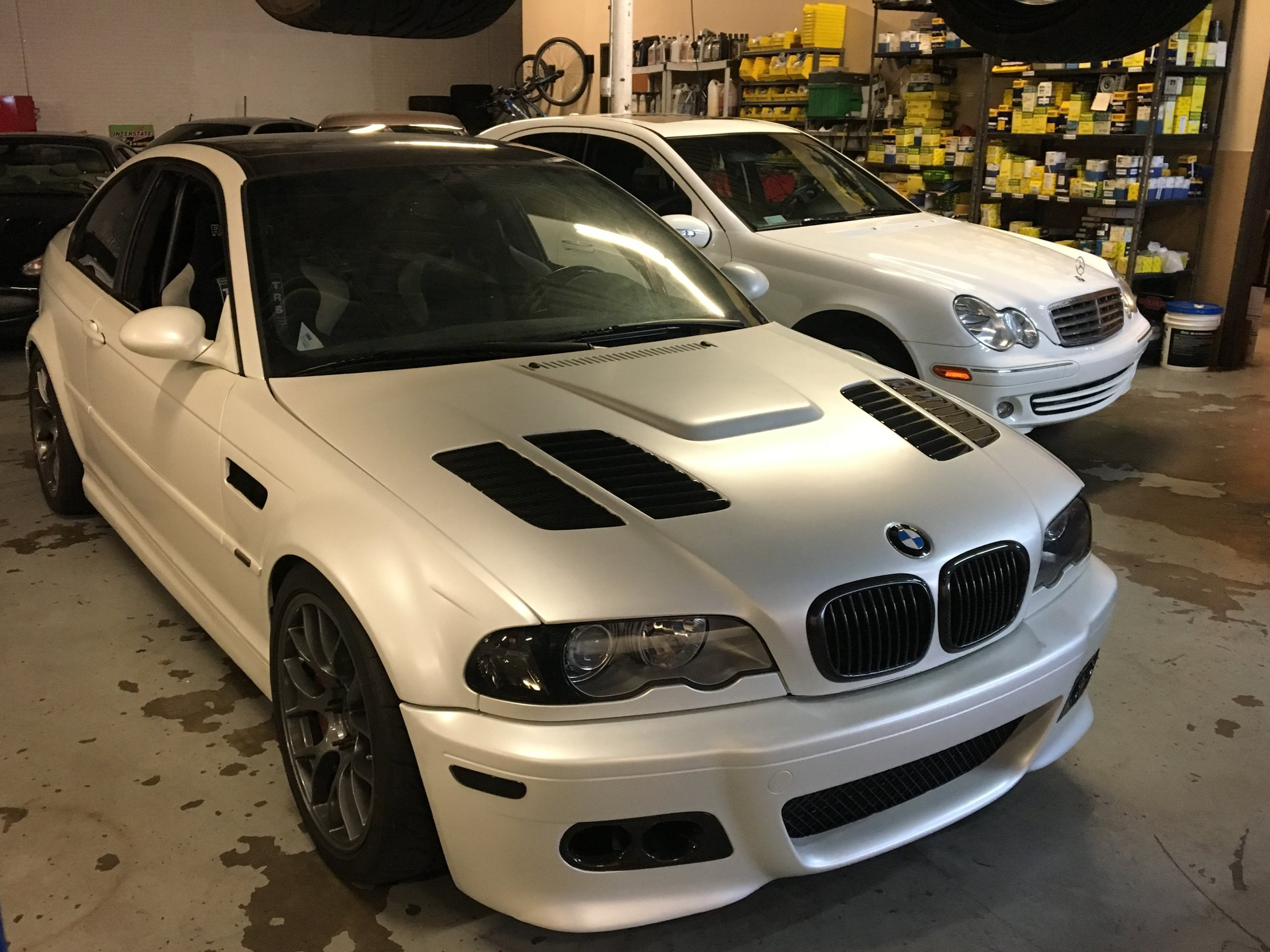 A white BMW E46 M3 sports car with black accents, parked indoors between other white Mercedes-Benz vehicles in a garage with shelves filled with automotive parts.