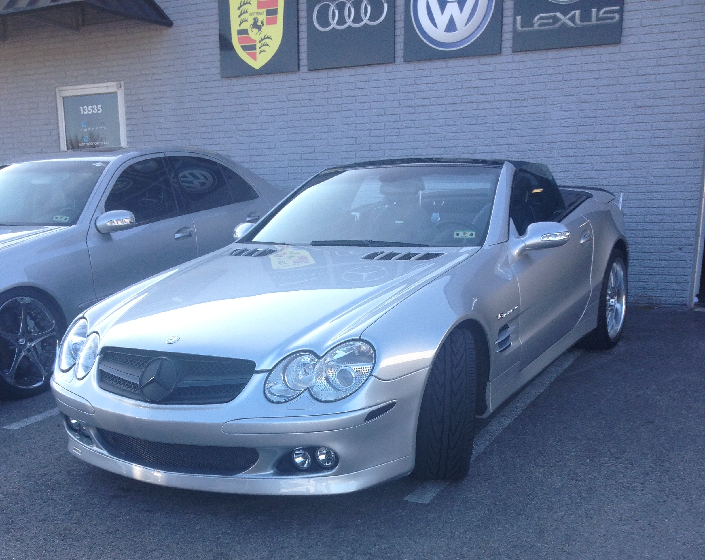 A silver Mercedes-Benz convertible sports car parked in front of a building with Porsche, Audi, Volkswagen, and Lexus signs.