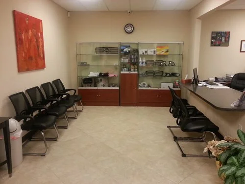 Empty waiting room with black chairs, glass display case with various glasses and items, a reception desk with a computer, and a few pieces of wall art
