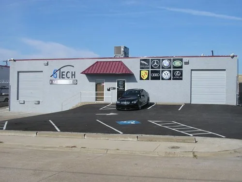Automotive repair shop with signs for various car brands, parking spaces including a handicapped spot, and a black car parked in front.