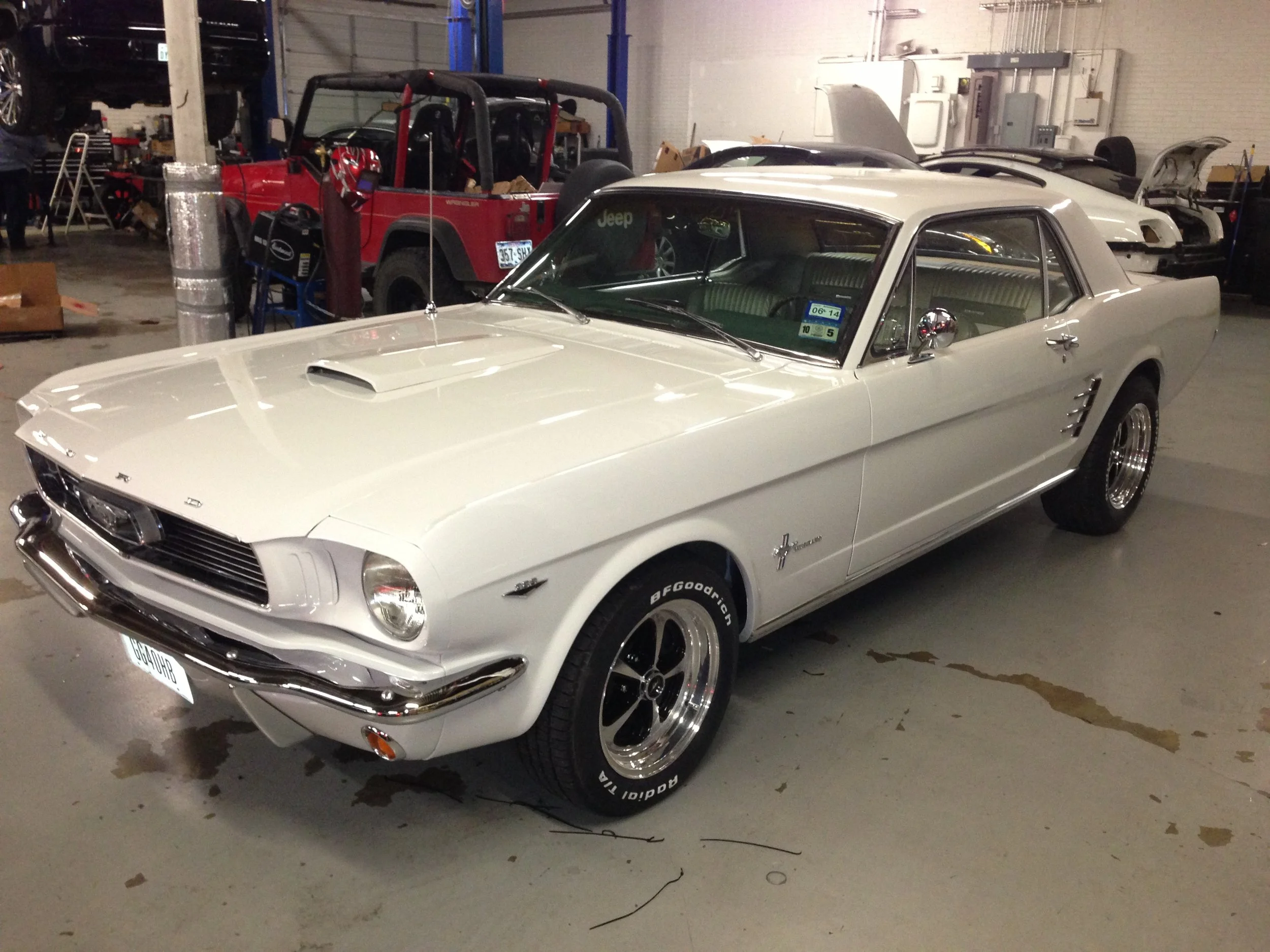 A vintage white Ford Mustang car parked indoors, with a hood scoop and chrome details, surrounded by other vehicles in the background.
