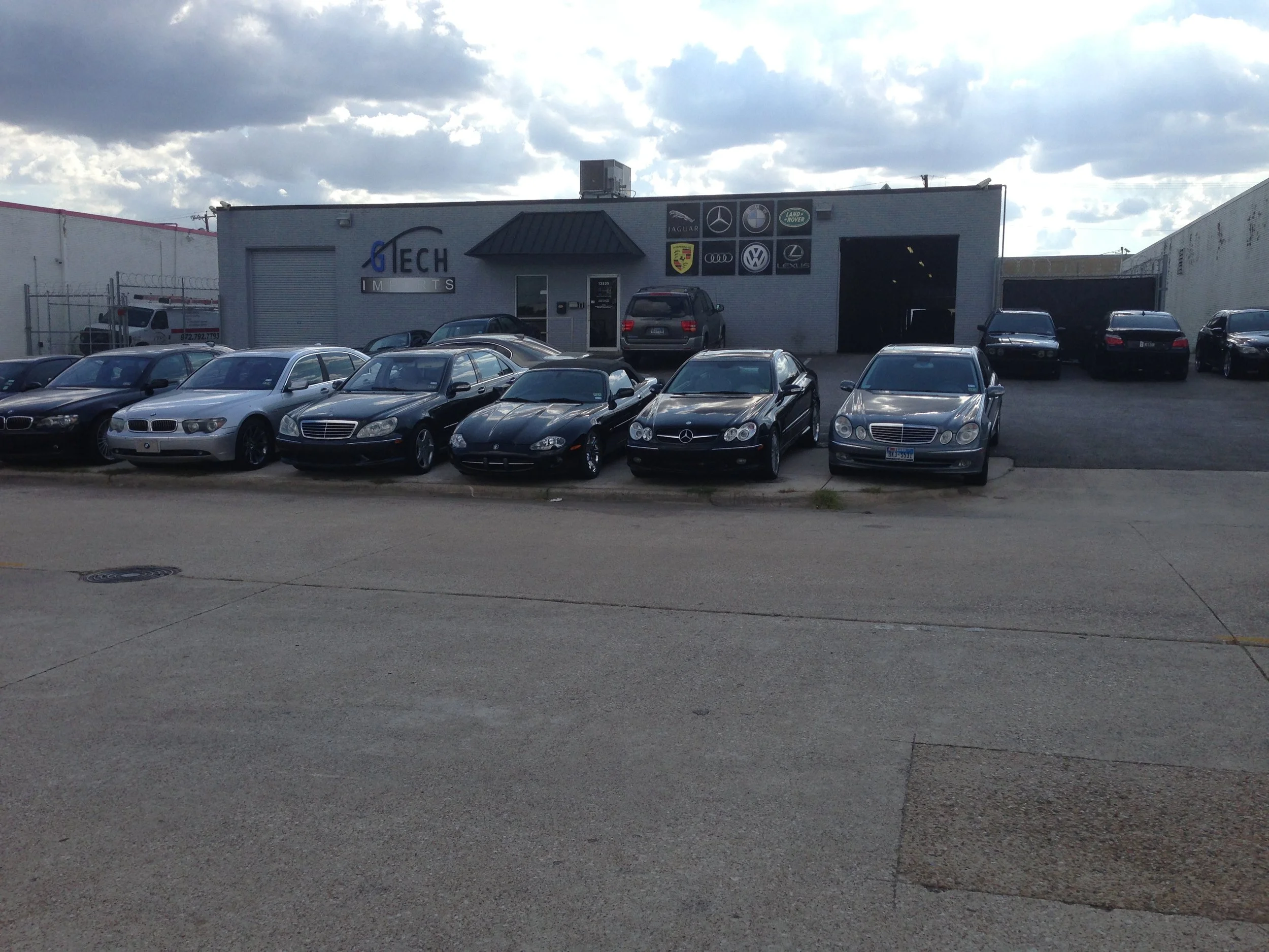 Car dealership lot with various luxury vehicles, including Mercedes-Benz and BMW, parked in front of a building labeled TECH Imports, with logos of luxury car brands on the building and a partly cloudy sky above.