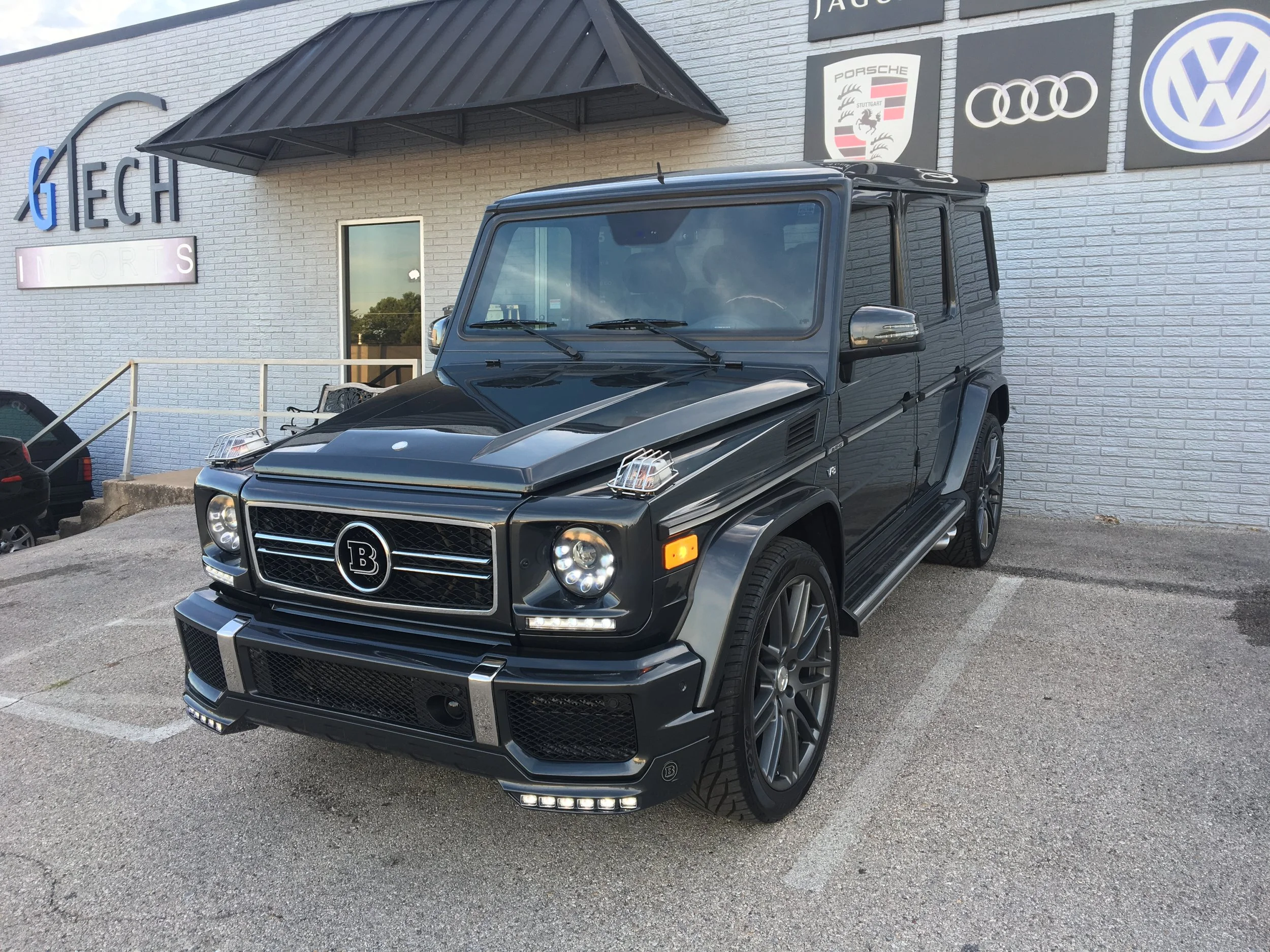 Black luxury SUV parked outside a building with car brand logos on the wall, including Porsche, Audi, Volkswagen, and Bentley.